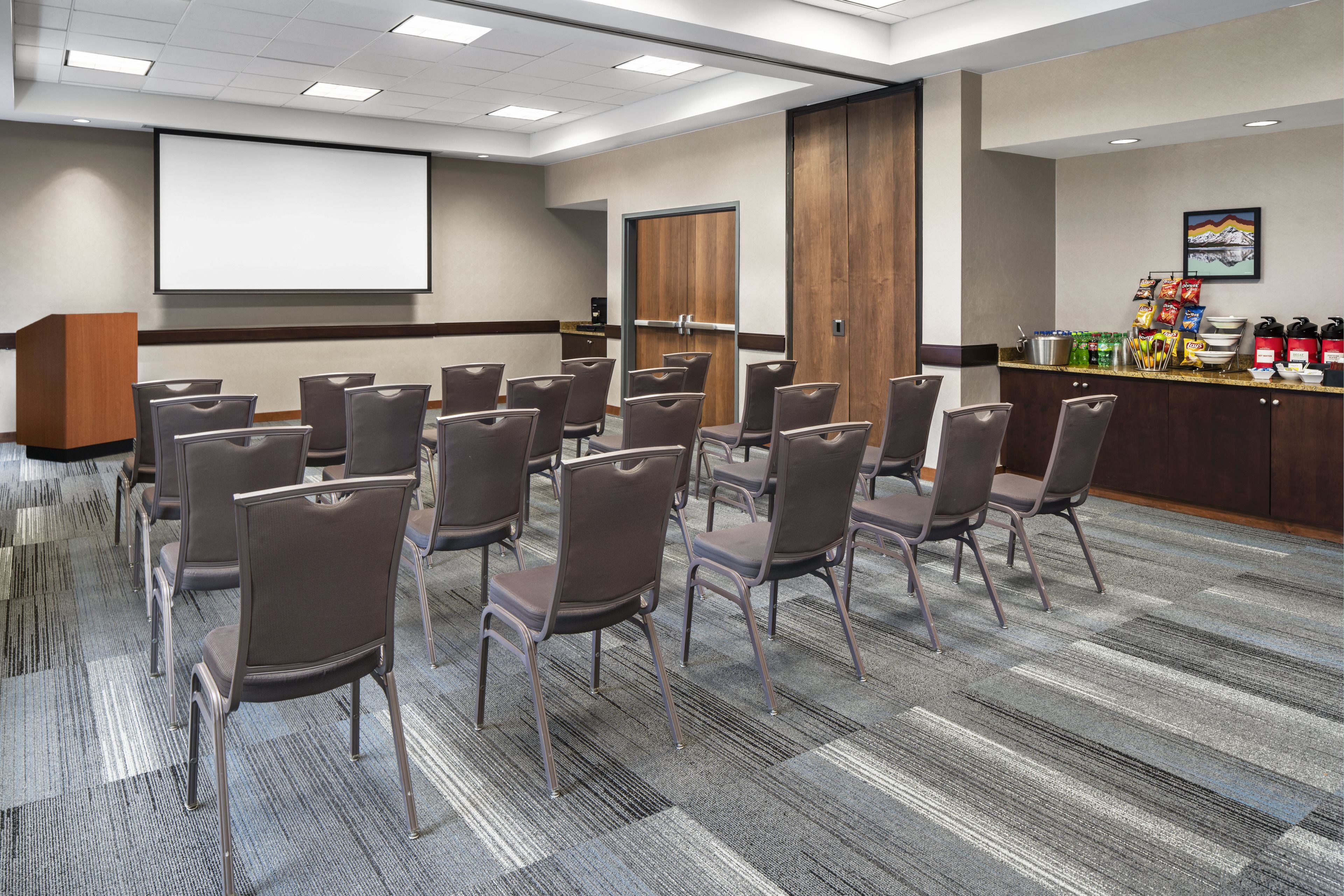 A conference room with rows of chairs, a projector screen, a wooden door, and a snack bar with various items.