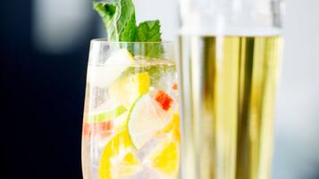 A hand holding a glass with a cocktail, garnished with lime and mint, and a tall glass of champagne in the background.