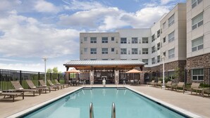 Una piscina con lettini e ombrelloni, circondata da un edificio a più piani e un’area patio coperta.