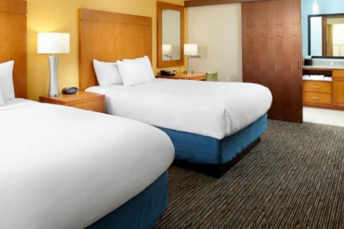 A hotel room with two beds, a wooden headboard, a nightstand with a lamp, a mirror, and a bathroom with a sink and mirror.