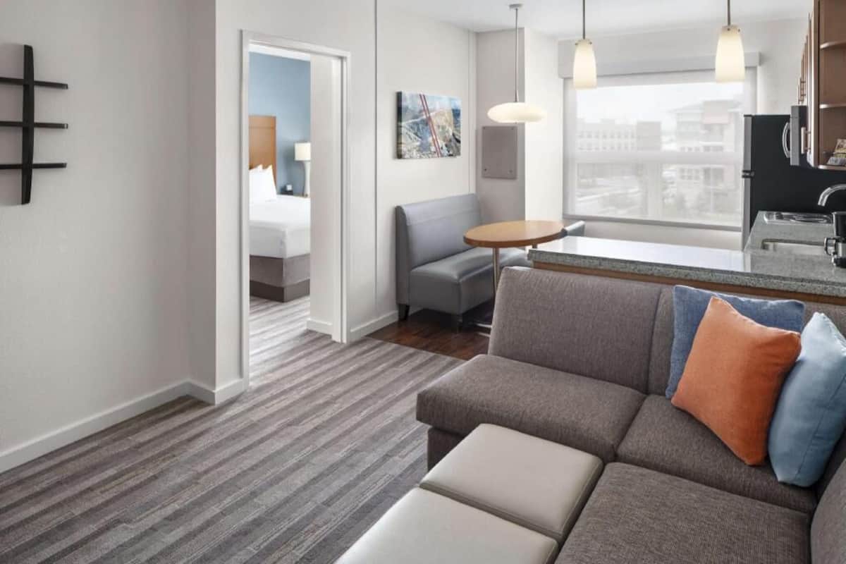 A modern hotel room with a gray sofa, a small round table, a city view through the window, and a kitchenette.