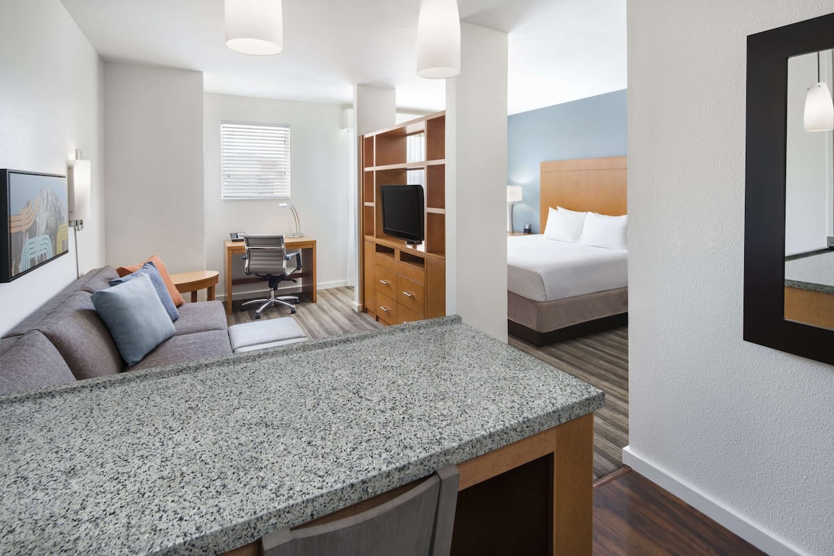 A hotel room with a kitchenette, a bed, a desk with a chair, a television, and a large mirror.