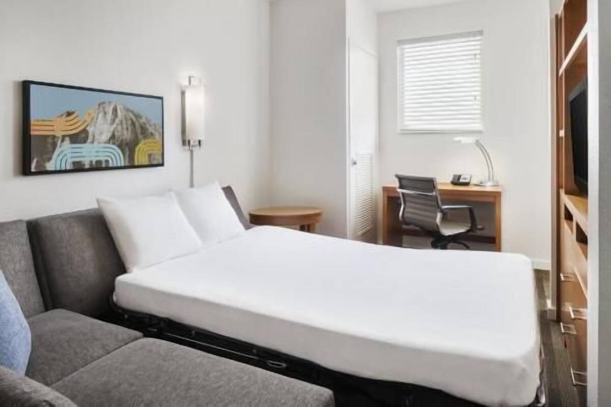 A hotel room with a bed, a desk with a chair, a small table, a lamp, and a painting on the wall.