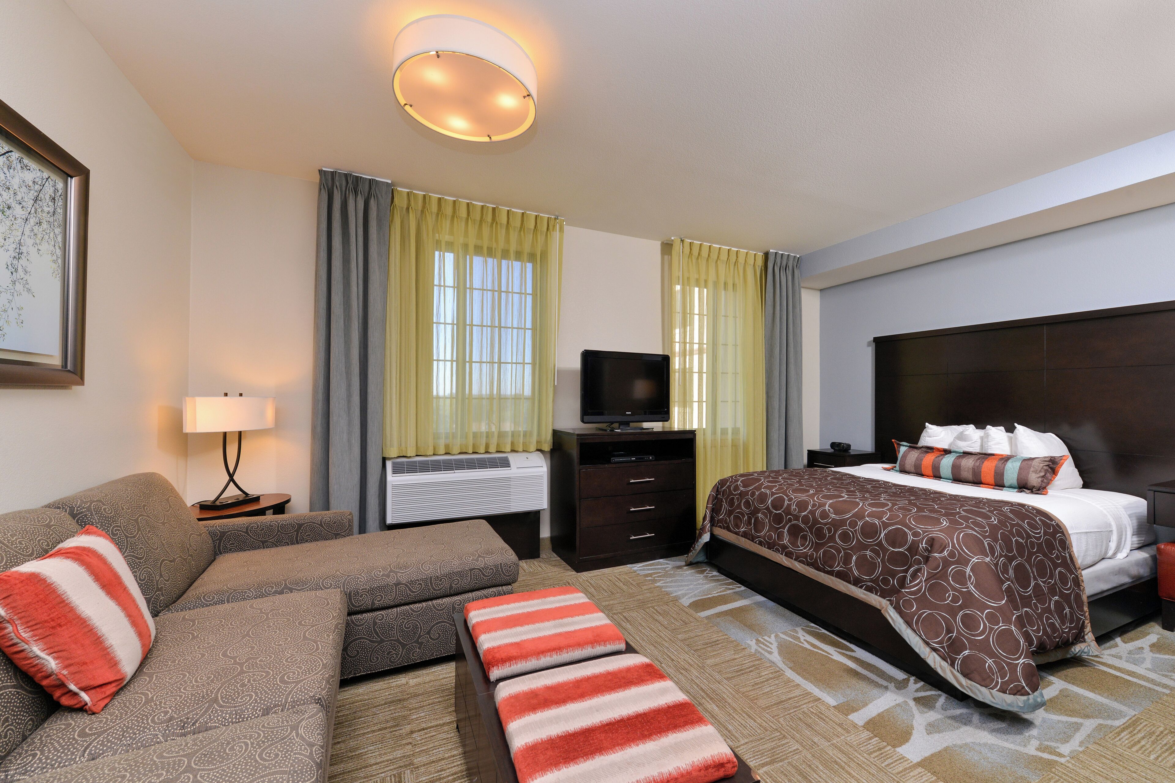 A hotel room with a sofa, a bed with a patterned bedspread, a flat-screen TV, a desk with a lamp, and a window with curtains.