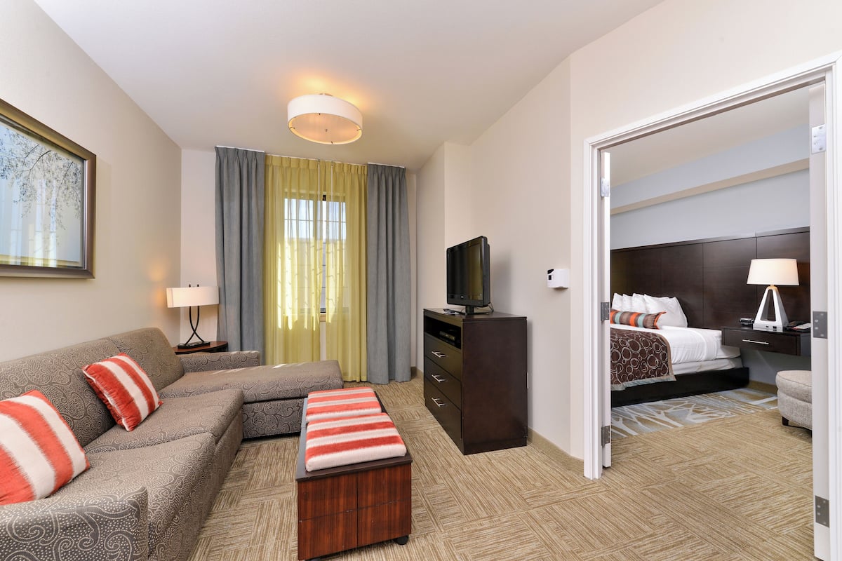 A hotel room with a sofa, ottoman, television, and a bed with a headboard.