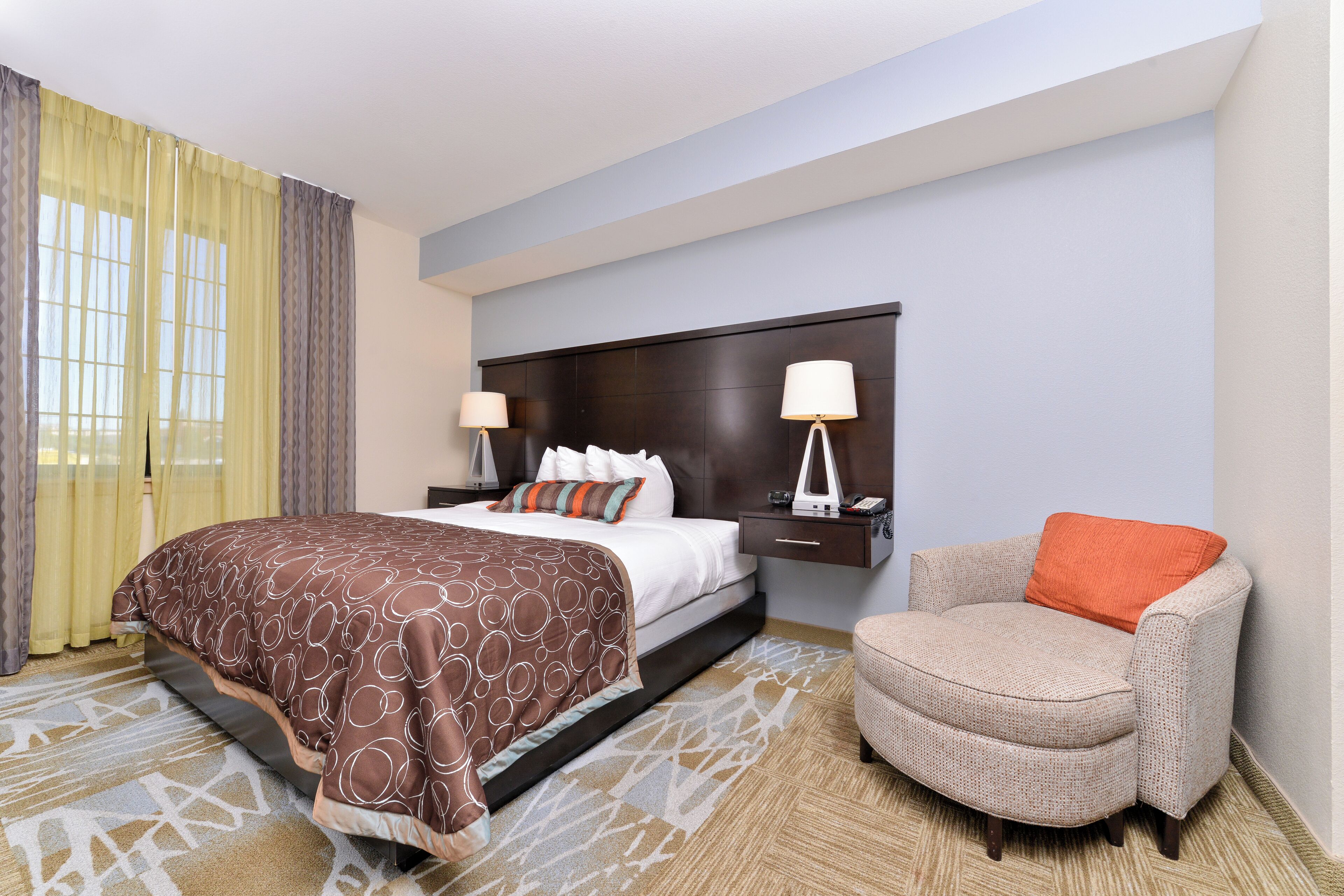 A hotel room with a bed, nightstands, a chair, and a large window with curtains.