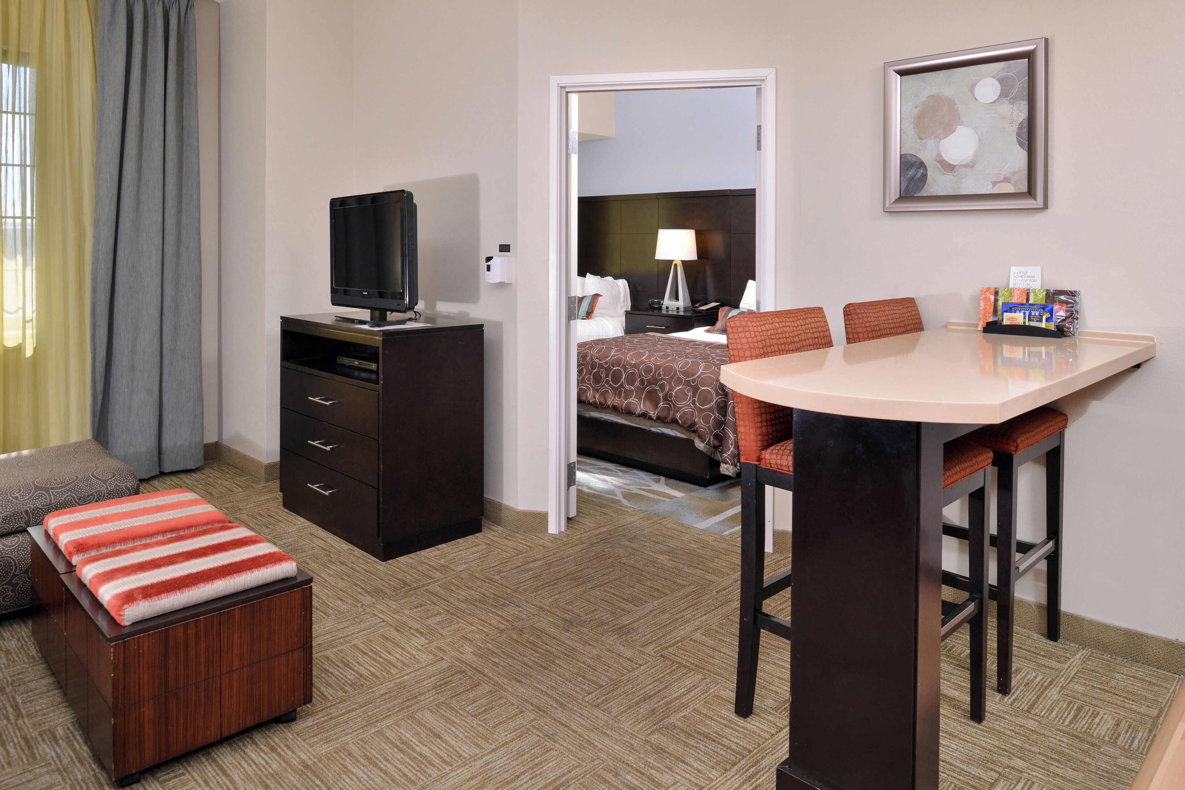 Une chambre d’hôtel comprenant un lit, un bureau avec une chaise, une télévision, une petite table avec une lampe et un tableau accroché au mur.