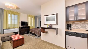 Ein Hotelzimmer mit Bett, einer Couch, einem kleinen Tisch, einem Fernseher, einer Küchenzeile und einem Fenster mit Vorhängen.