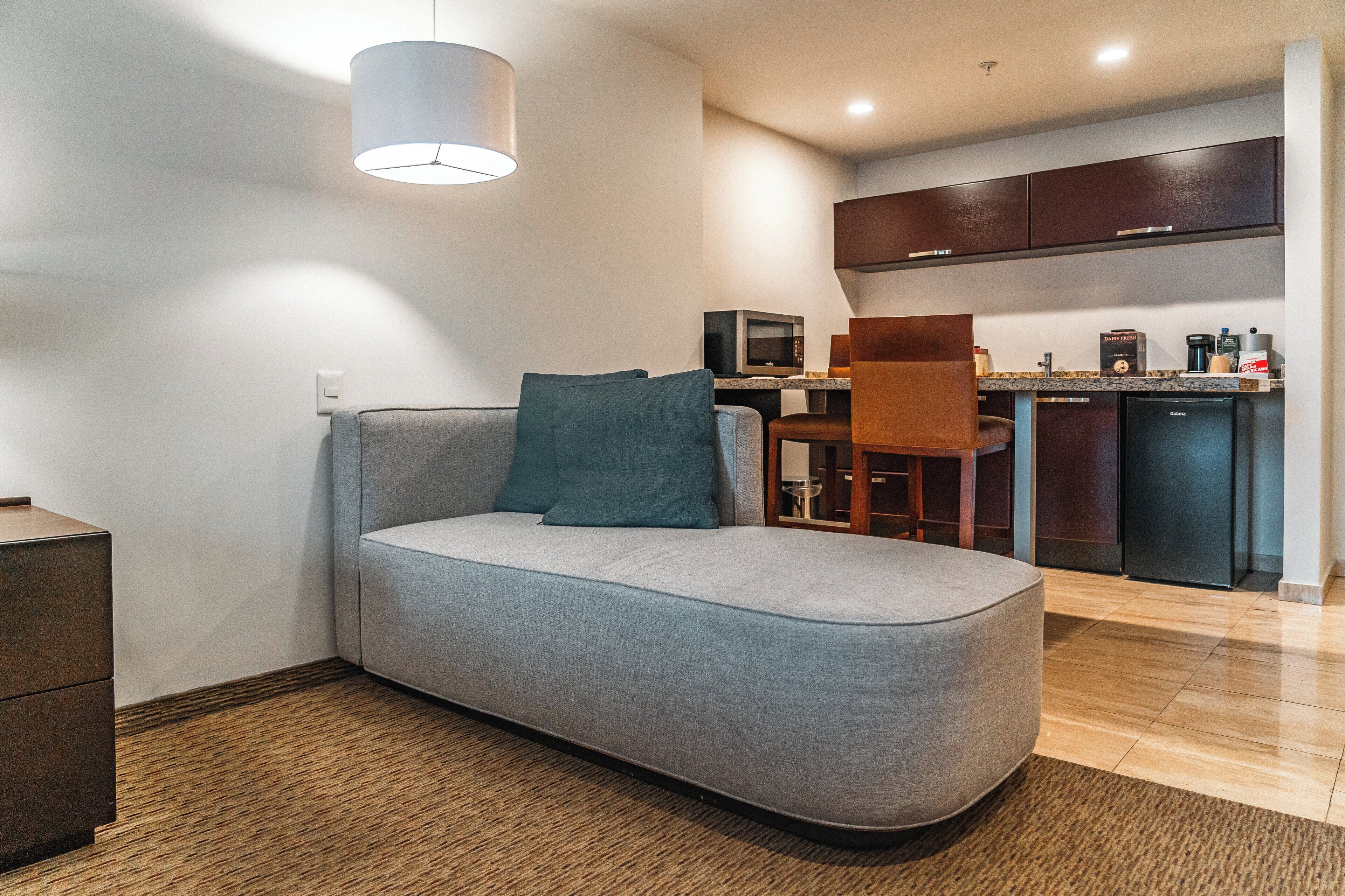 A modern living room with a grey sofa, a wooden coffee table, and a dining area with a table and chairs. A kitchen with dark cabinets and a refrigerator is visible in the background.