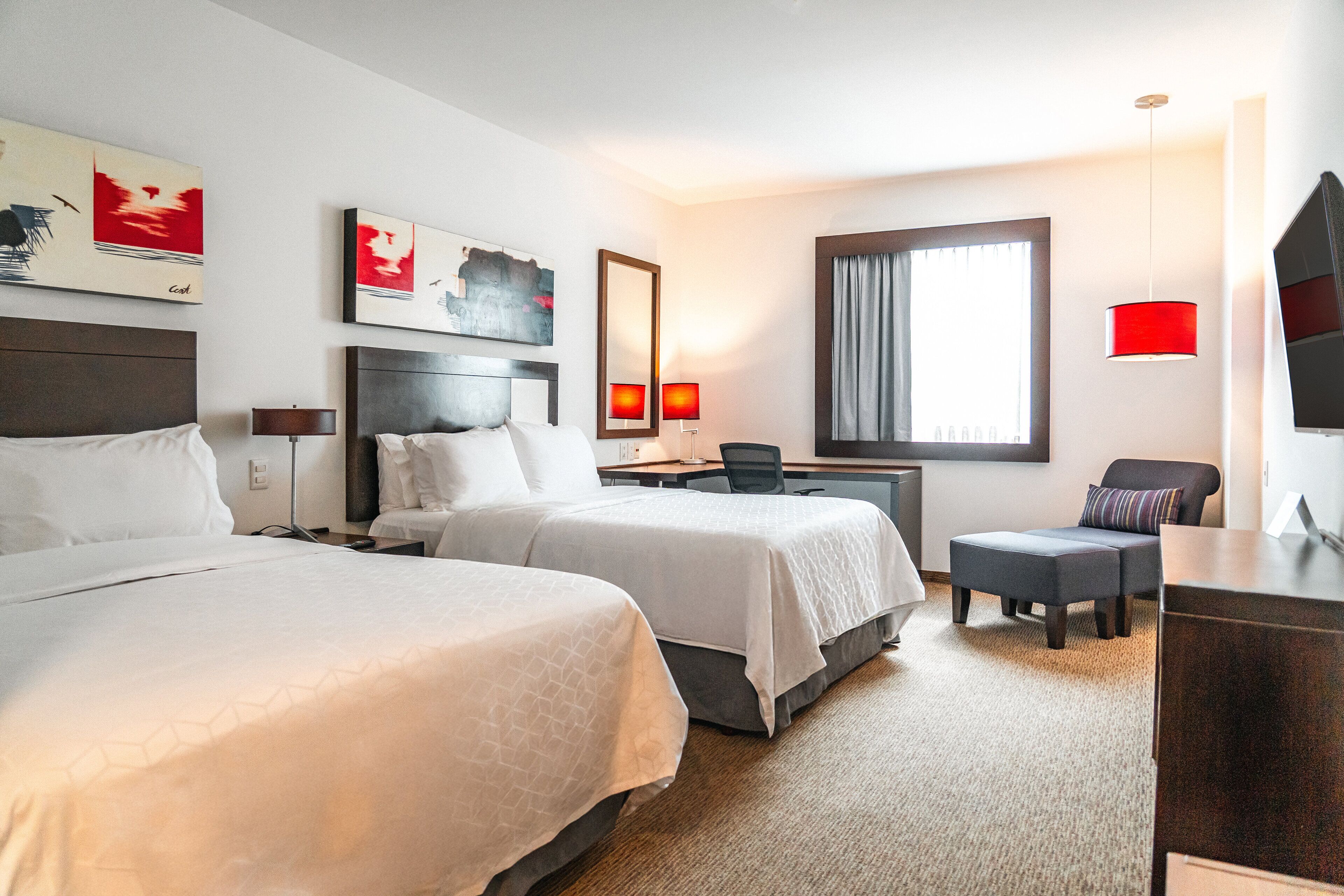 A hotel room with two beds, a desk, a chair, a TV, and artwork on the walls.