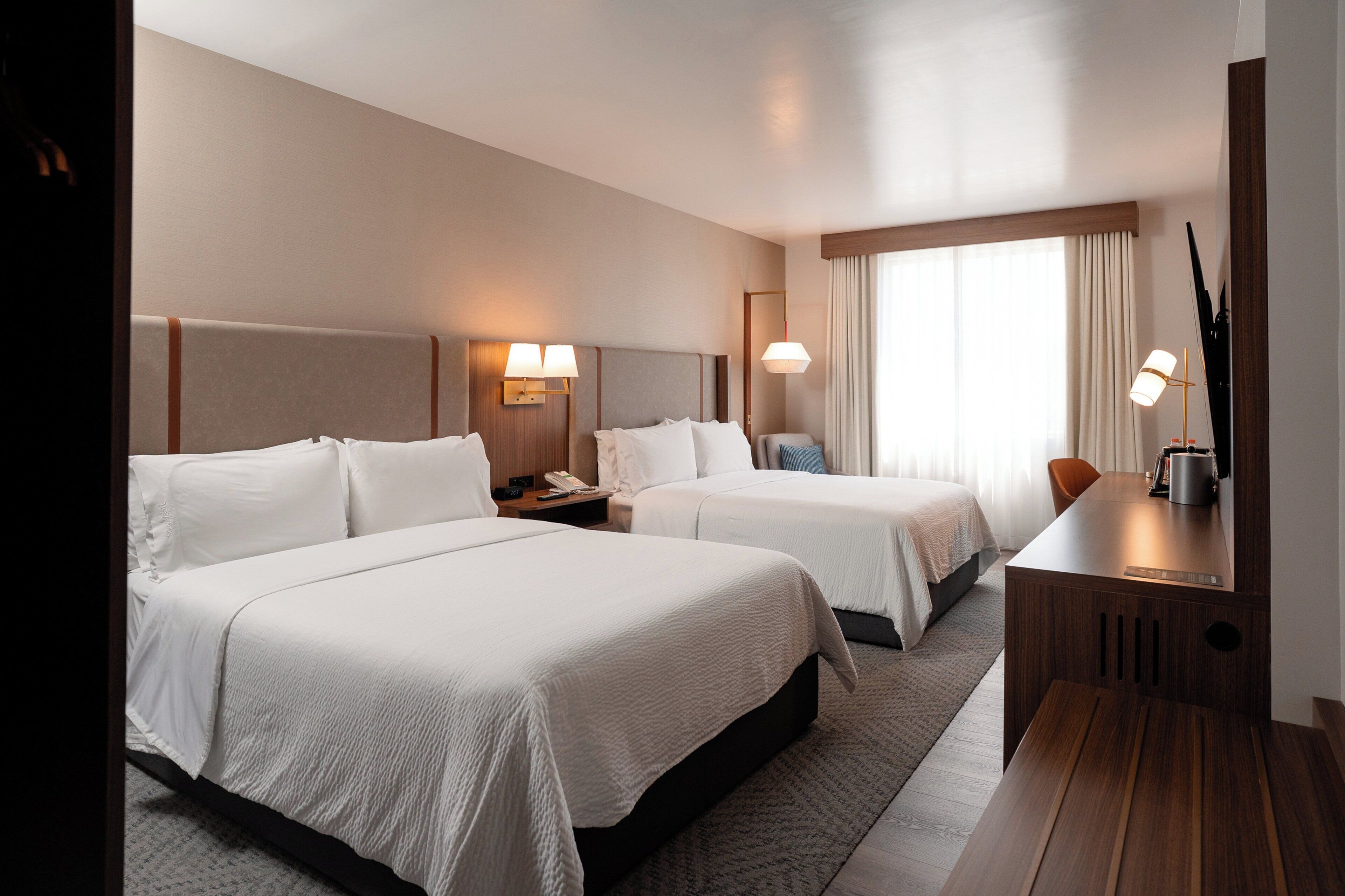 A hotel room with two beds, a desk, a chair, a lamp, and a large window with curtains.