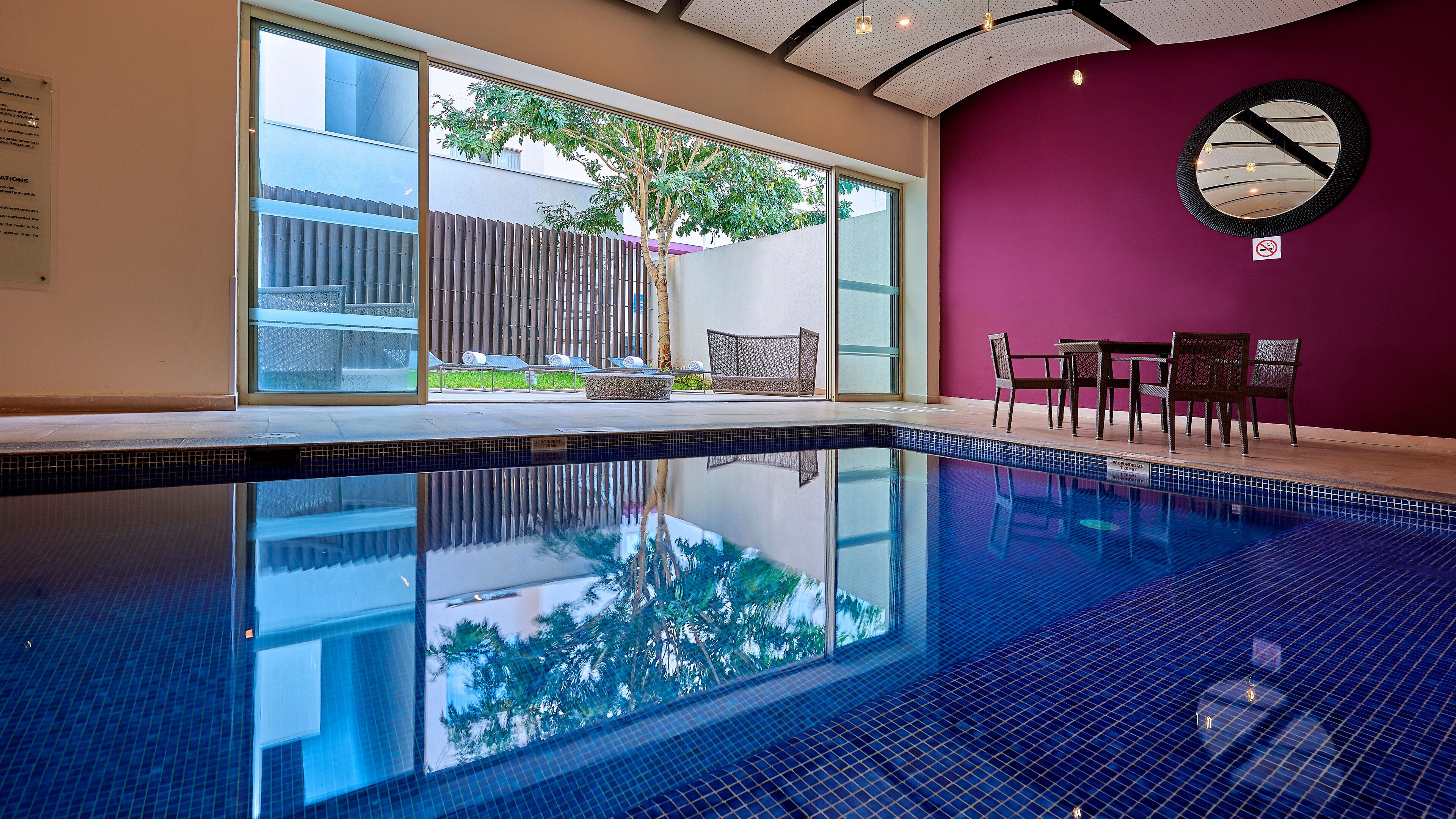 A swimming pool with a clear view of the outside, a seating area with chairs, and a wall-mounted clock.