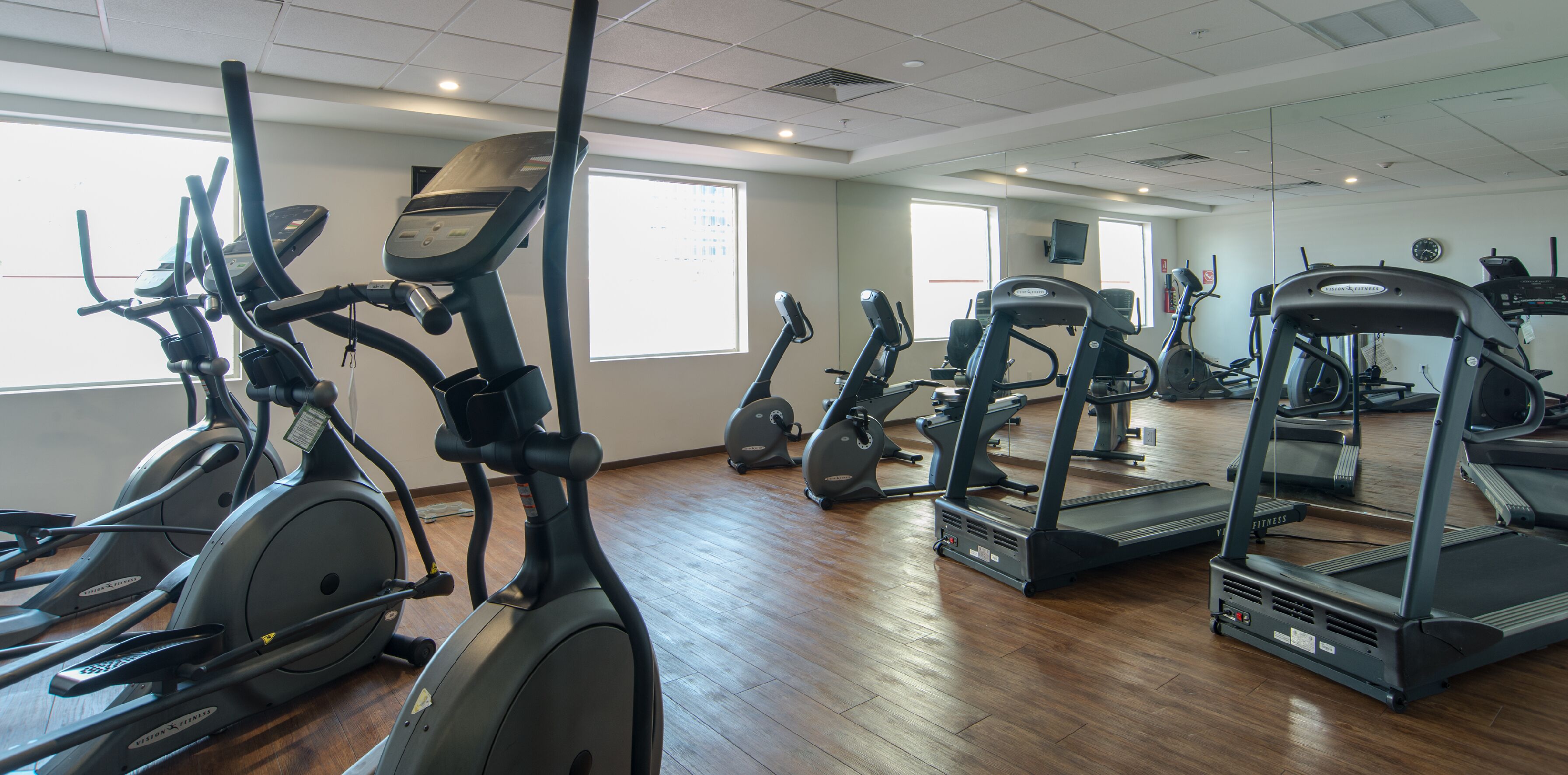 A gym with various cardio machines, large windows, and a wooden floor.