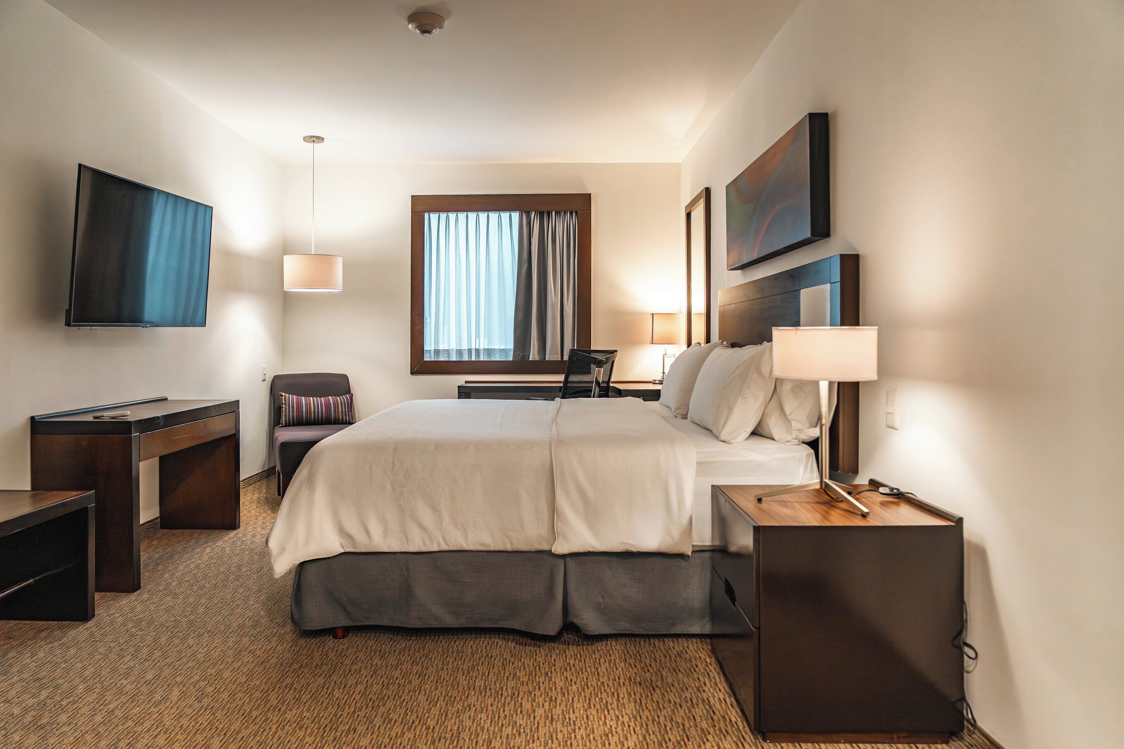 A hotel room with a bed, bedside tables, a desk, a TV, and a window with curtains.
