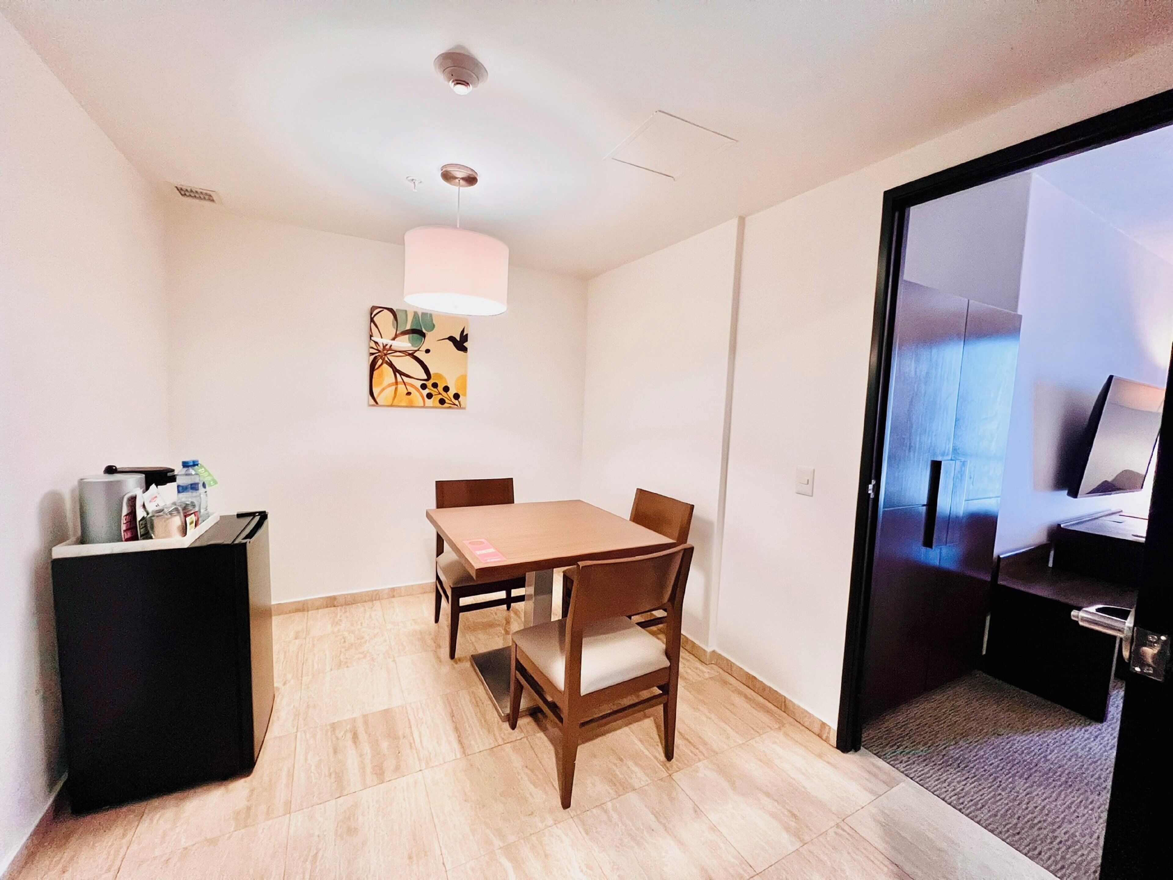 A hotel room with a dining table, chairs, a small black cabinet, a painting on the wall, and a sliding door leading to a bed.