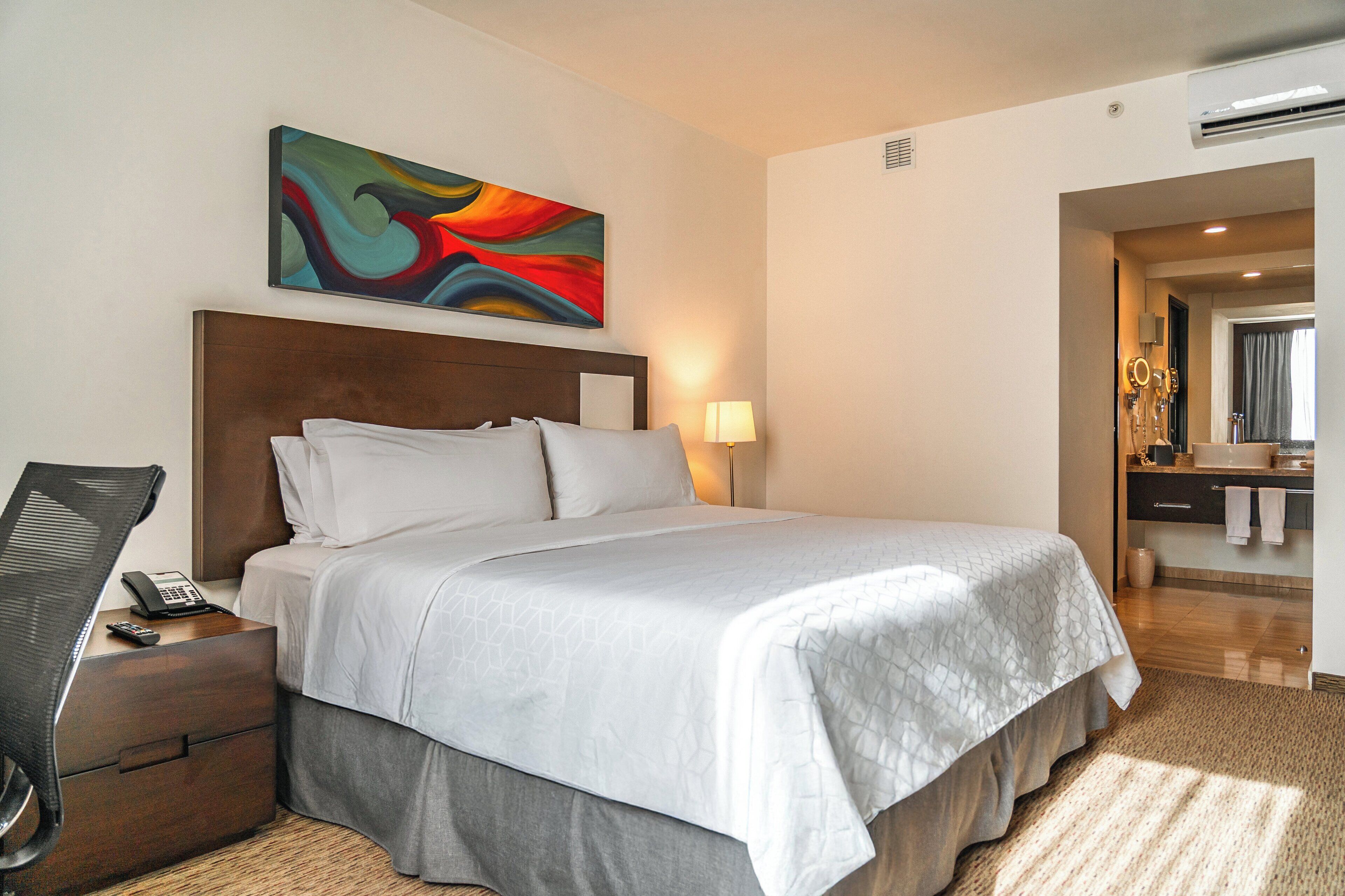 Een hotelkamer met een groot bed, een bureau met stoel, een telefoon en een kleurrijk abstract schilderij aan de muur.