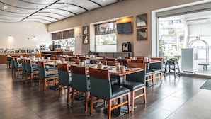 Un restaurante amplio con varias mesas y sillas, una estación de café y ventanas grandes que permiten la entrada de luz natural.