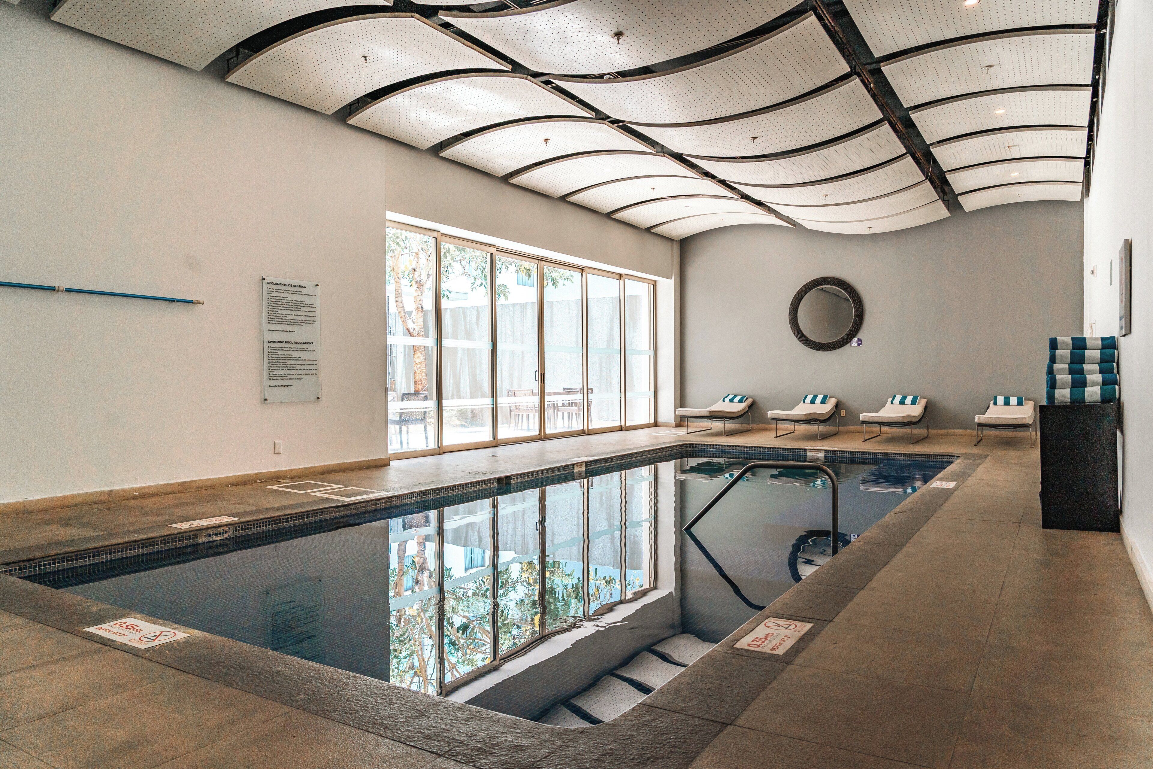 An indoor swimming pool with clear glass walls, a tiled floor, and a ceiling with a perforated design.