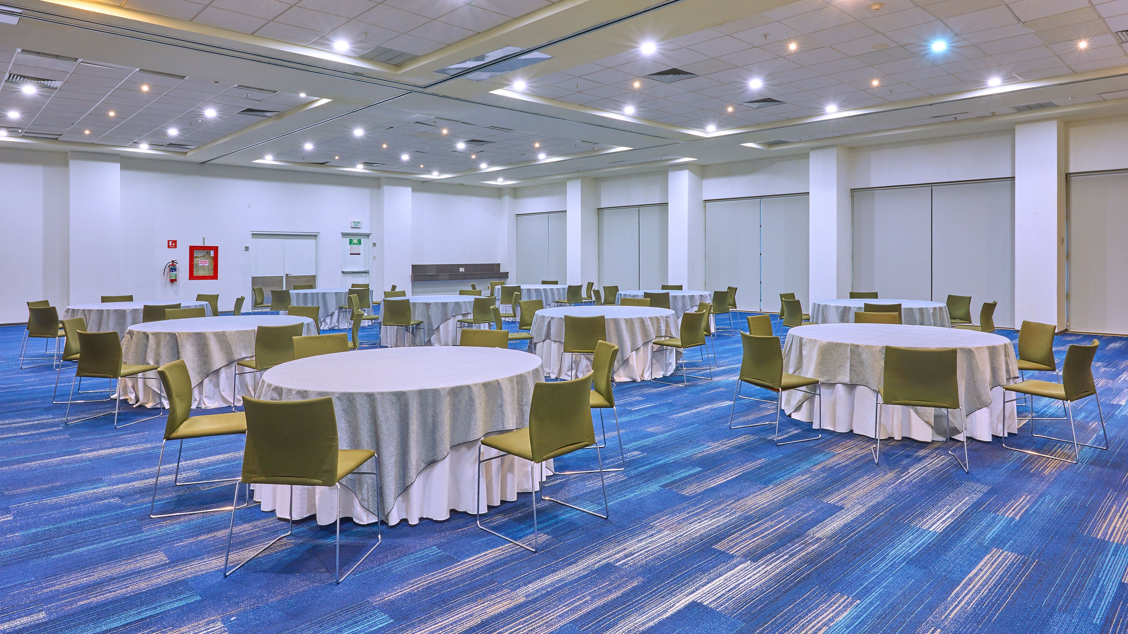 Una sala de conferencias con varias mesas redondas y sillas, piso alfombrado y techo blanco con iluminación.