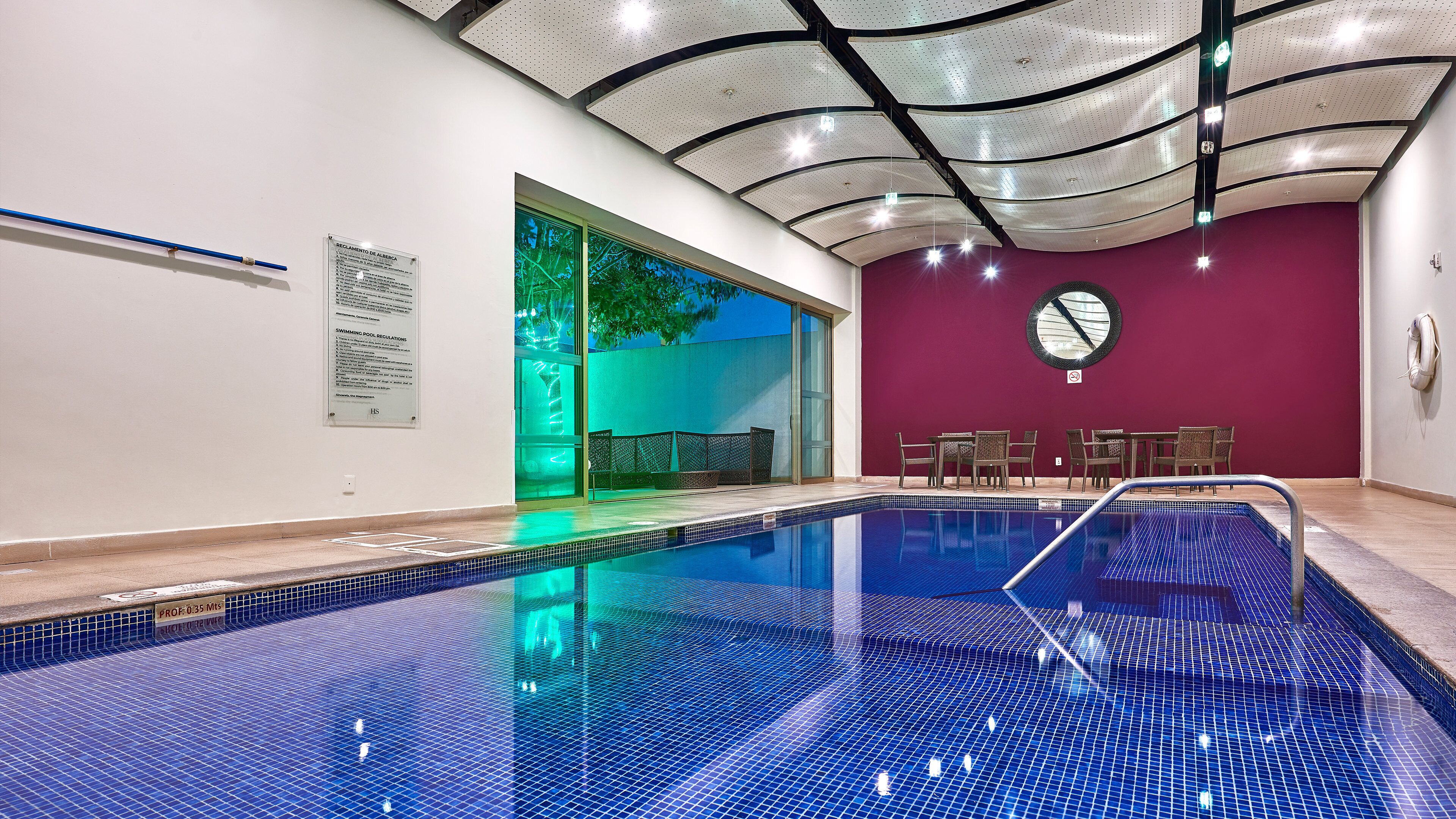 A swimming pool with a tiled floor and a ceiling with a unique design. There are chairs and a table in the background.
