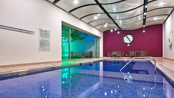Ein Schwimmbecken mit gefliestem Boden und einer Decke mit einem einzigartigen Design. Im Hintergrund befinden sich Stühle und ein Tisch.