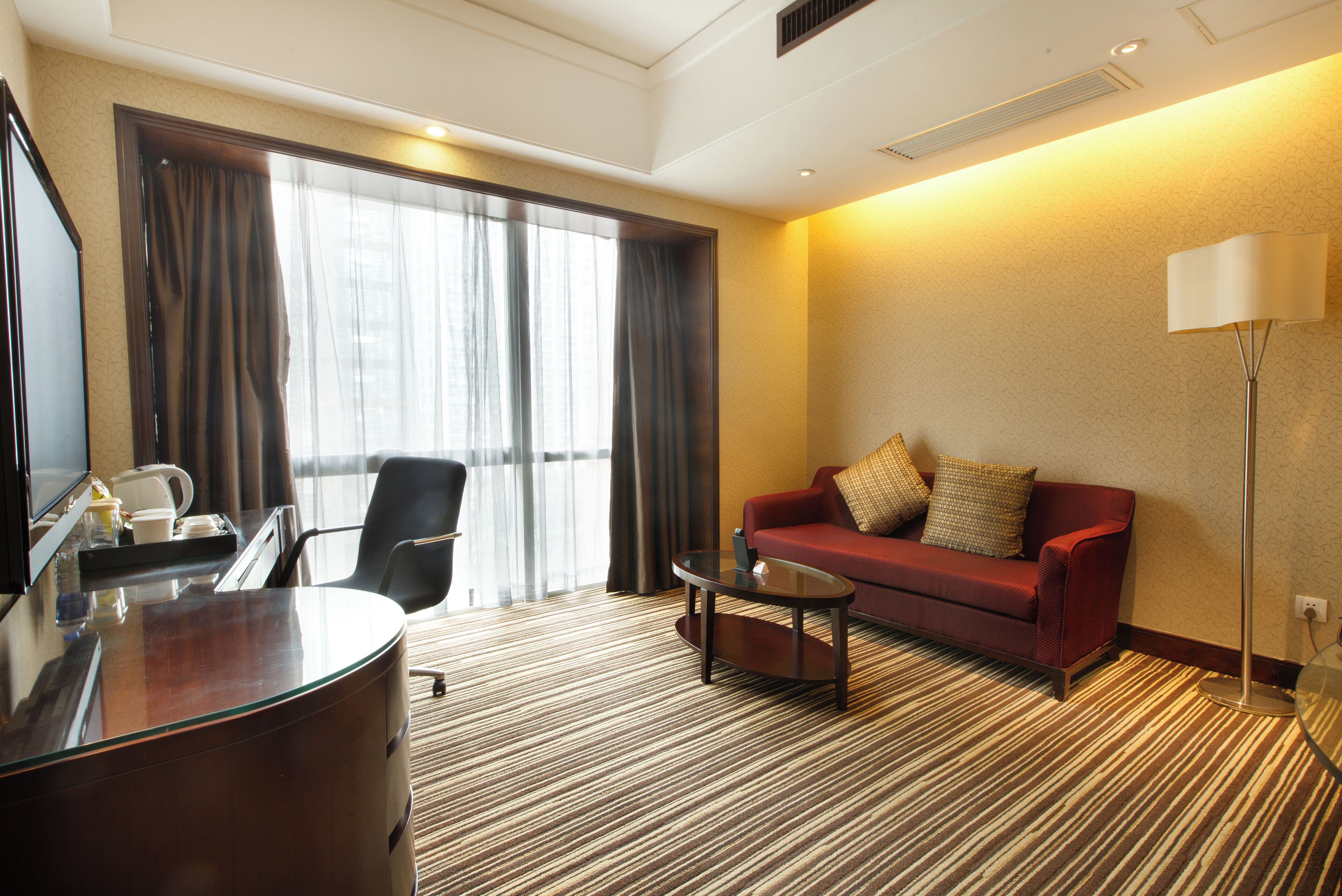 A hotel room with a desk, a red sofa, a glass coffee table, and a floor lamp.