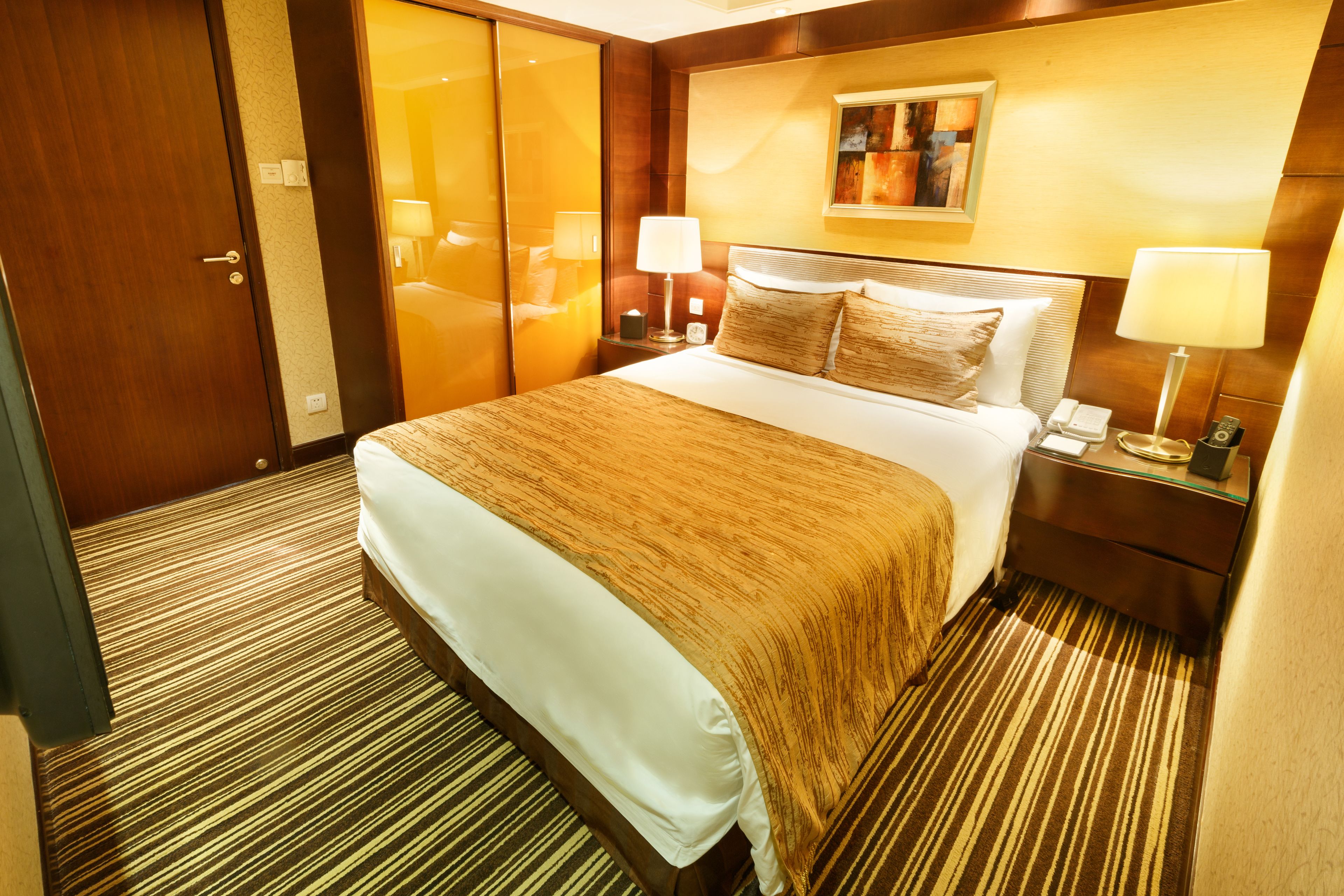 A hotel room with a large bed, a nightstand, a lamp, a painting on the wall, and a door.
