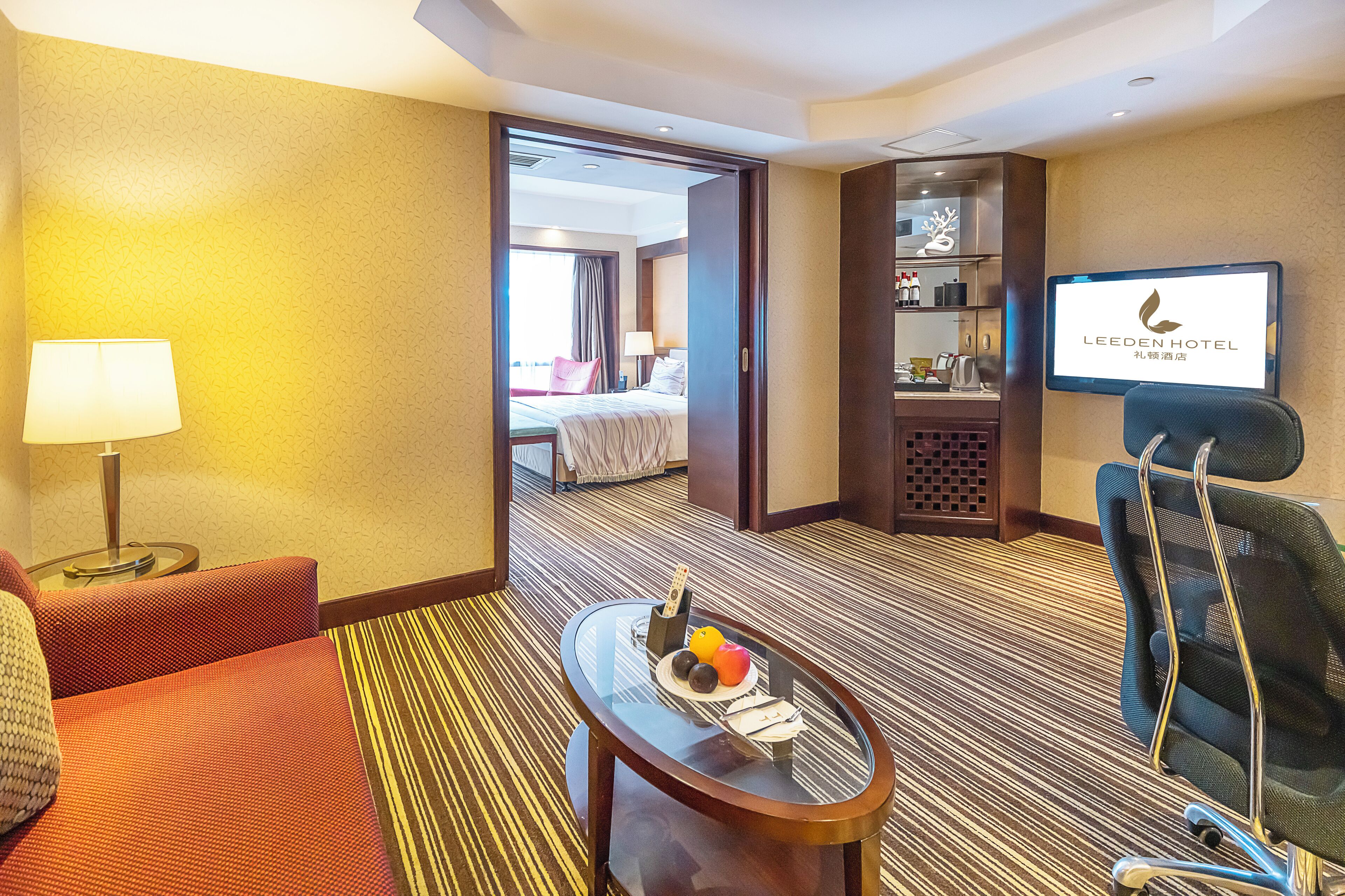 A hotel room with a bed, a TV, a desk with a lamp, a chair, and a small table with fruits and a remote control.