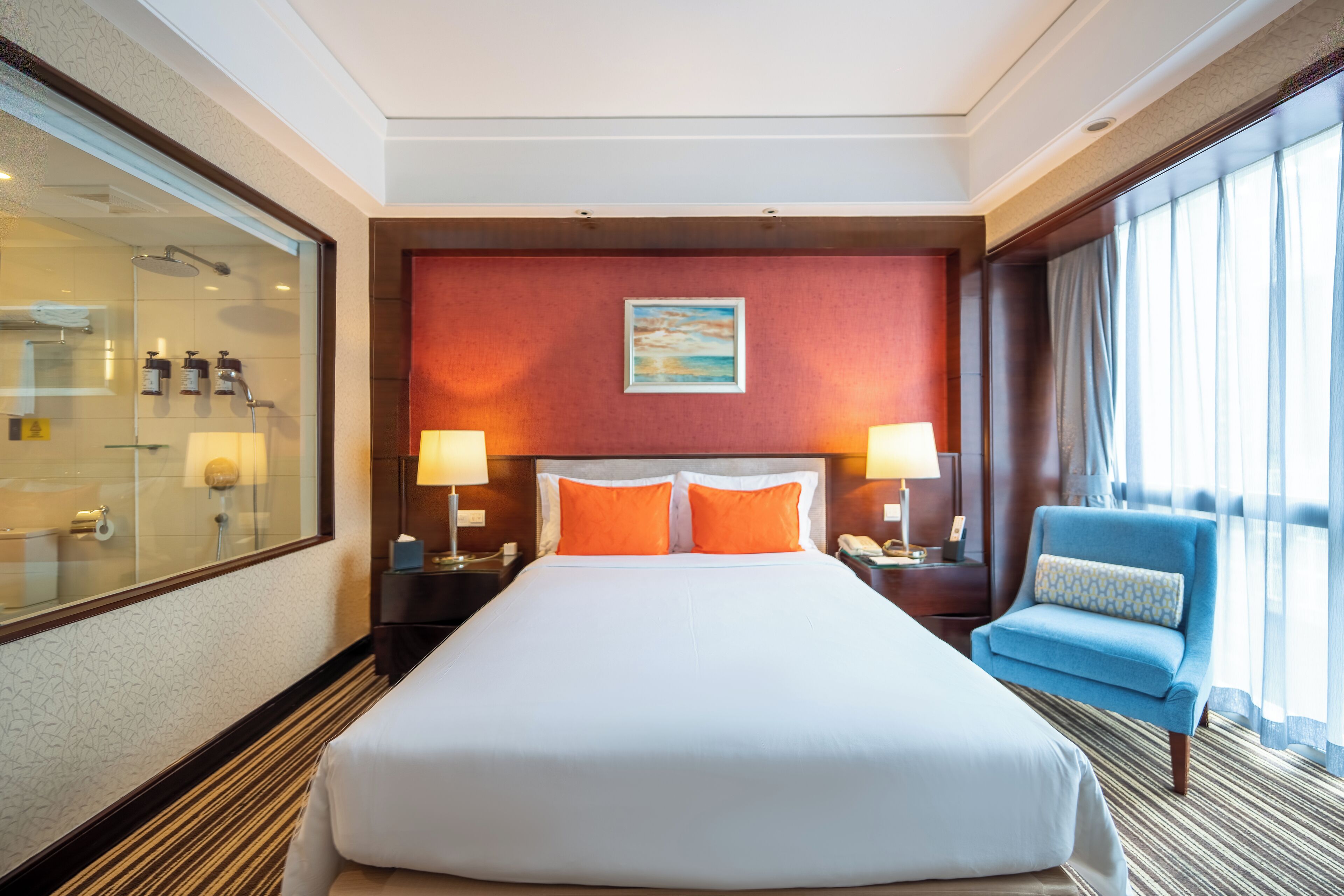 A hotel room with a large bed, two bedside lamps, a blue armchair, a painting on the wall, and a large window with curtains.