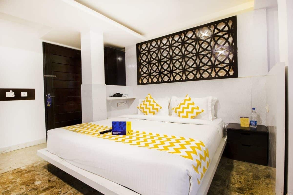 A modern hotel room with a large bed, a decorative wall panel, a nightstand, and a bathroom door.