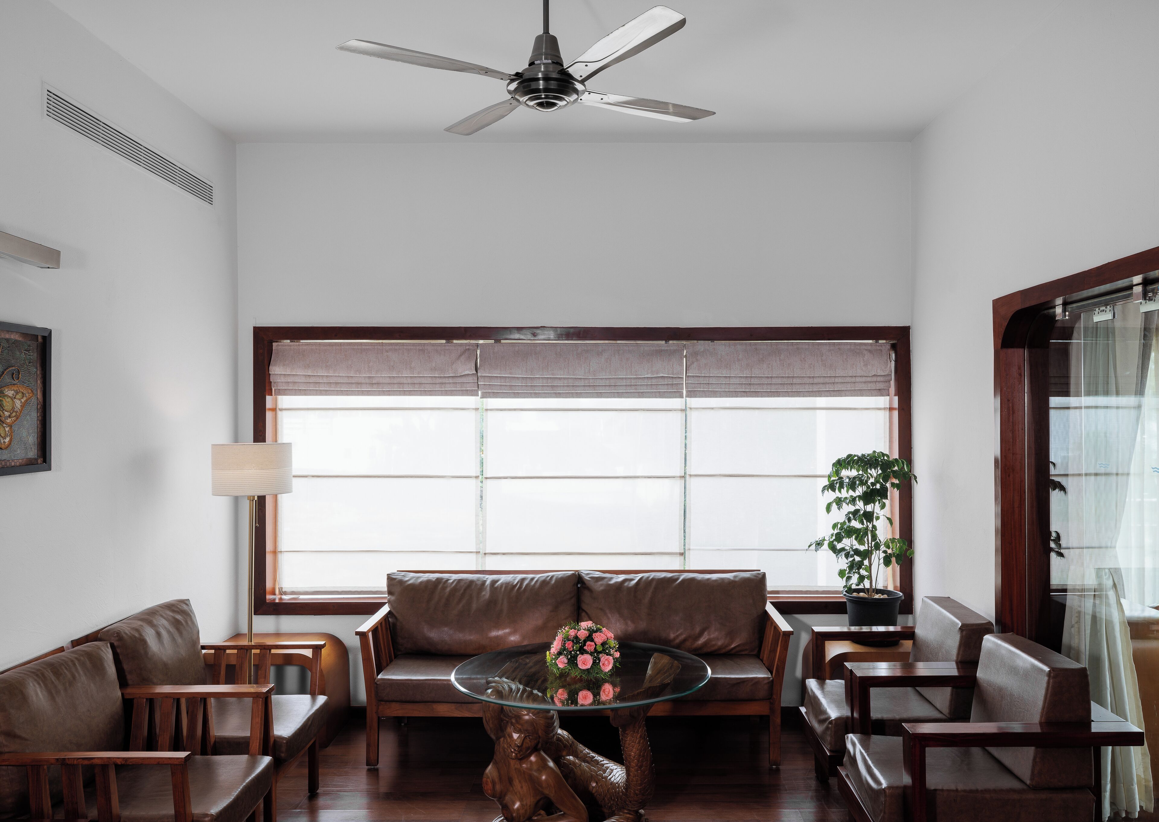 A modern living room with a brown leather sofa, wooden chairs, a glass coffee table with a floral centerpiece, a ceiling fan, and a large window with a view.