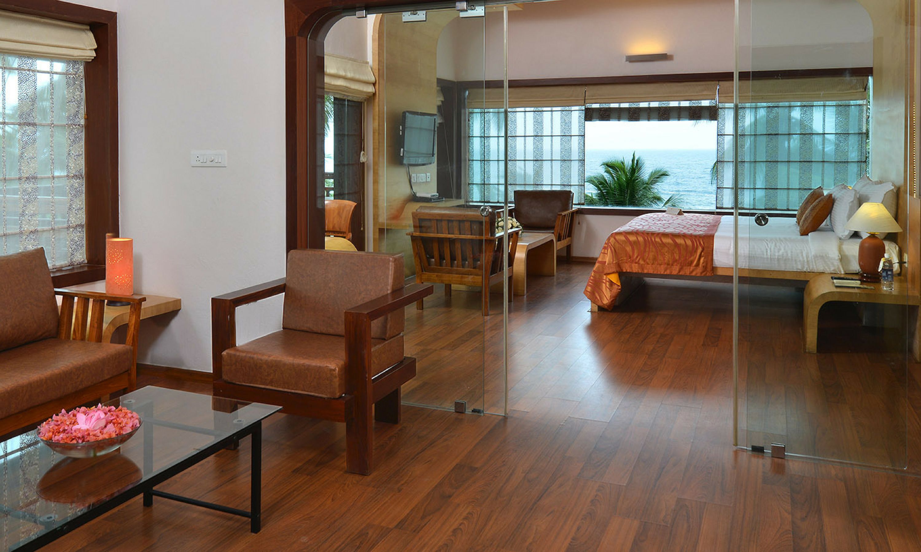 A hotel room with a wooden floor, a sofa, a chair, a bed, a nightstand, a lamp, and a window with a view of the sea.