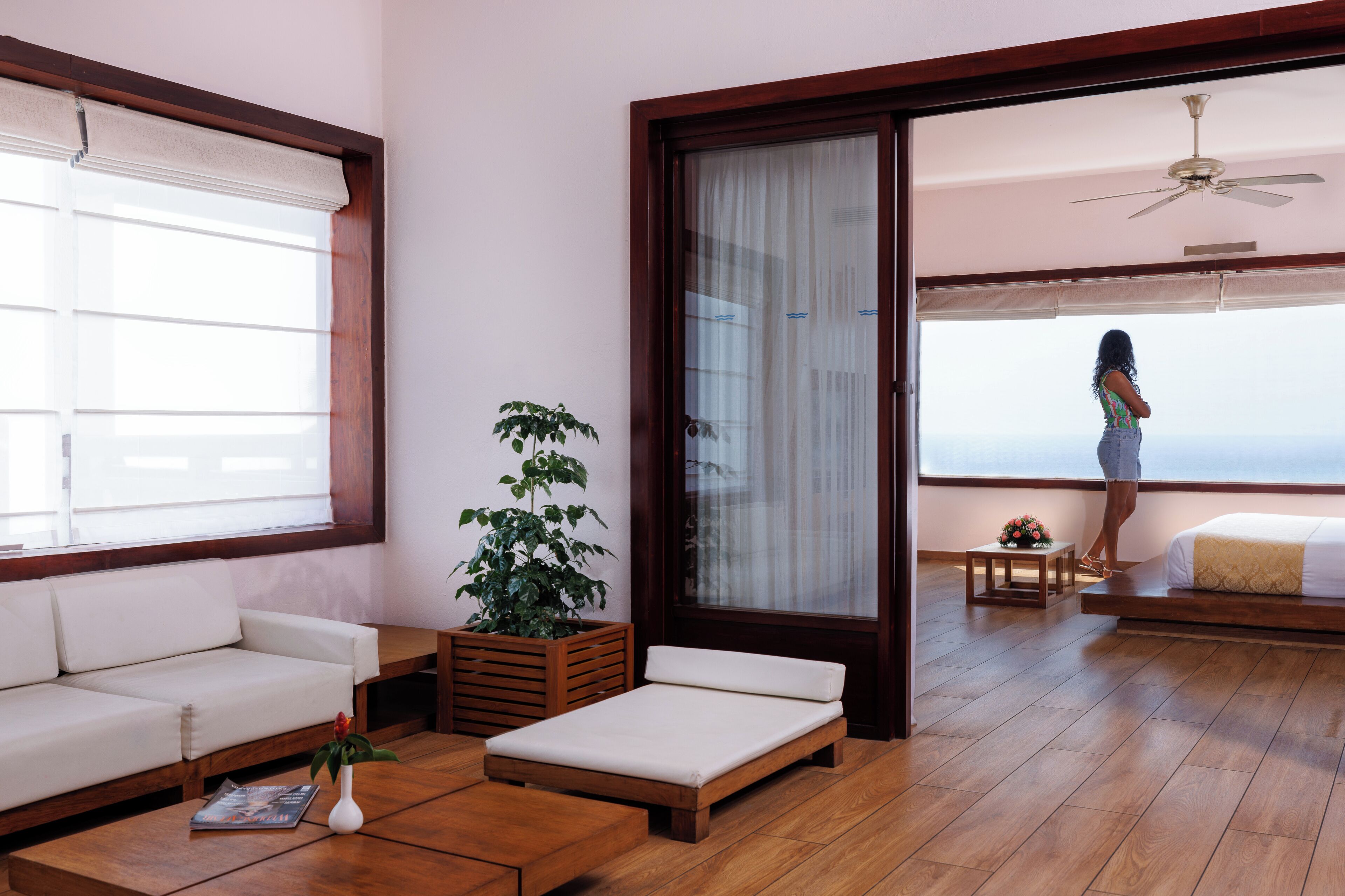 Una habitación con piso de madera, un sofá blanco, un banco de madera, una mesita con un florero y una planta, y vista al océano a través de una puerta corredera.