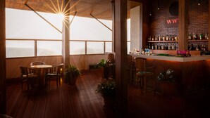 Zona de bar con suelo de madera, una barra y estantes con botellas. En primer plano, sillas y una mesa, y grandes ventanales con vistas al mar.