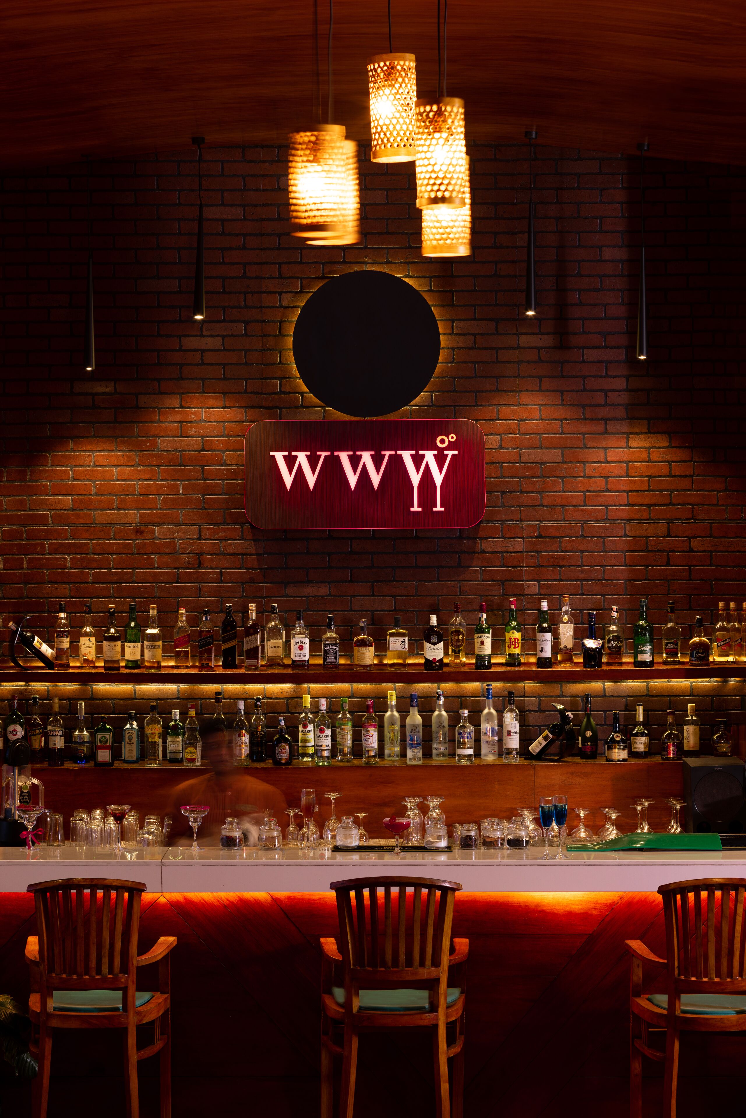 Eine Bar mit Backsteinwand, einem Schild mit 'WWYY' und verschiedenen Flaschen und Gläsern auf den Regalen.