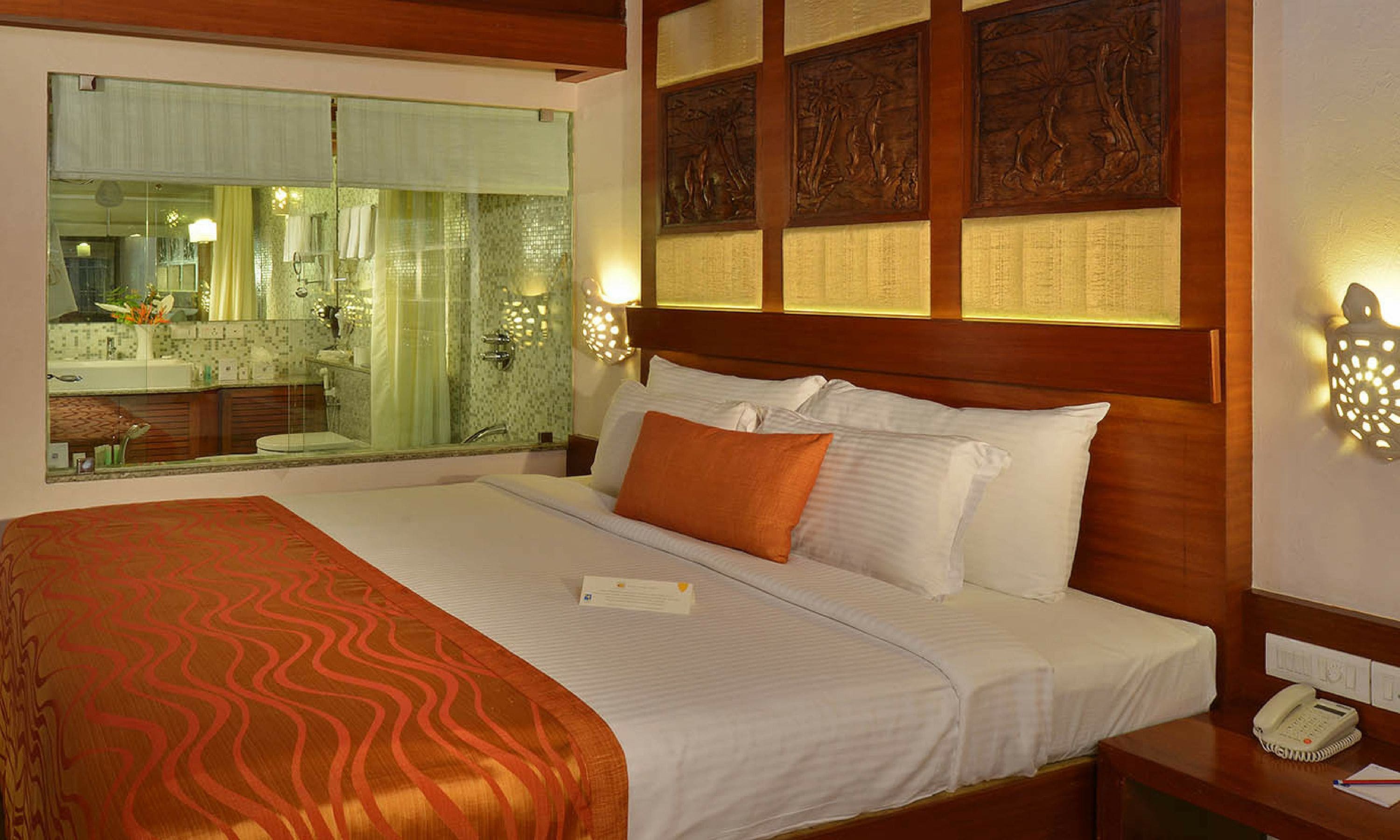 Habitación de hotel con una cama grande, un teléfono en la mesita de noche y vistas al baño a través de un separador de vidrio.