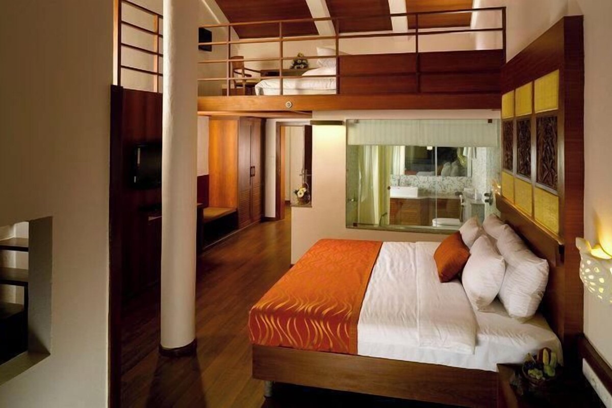 A bedroom with a bunk bed, a large bed with white bedding, a wooden floor, a glass door leading to a bathroom, and a small seating area.