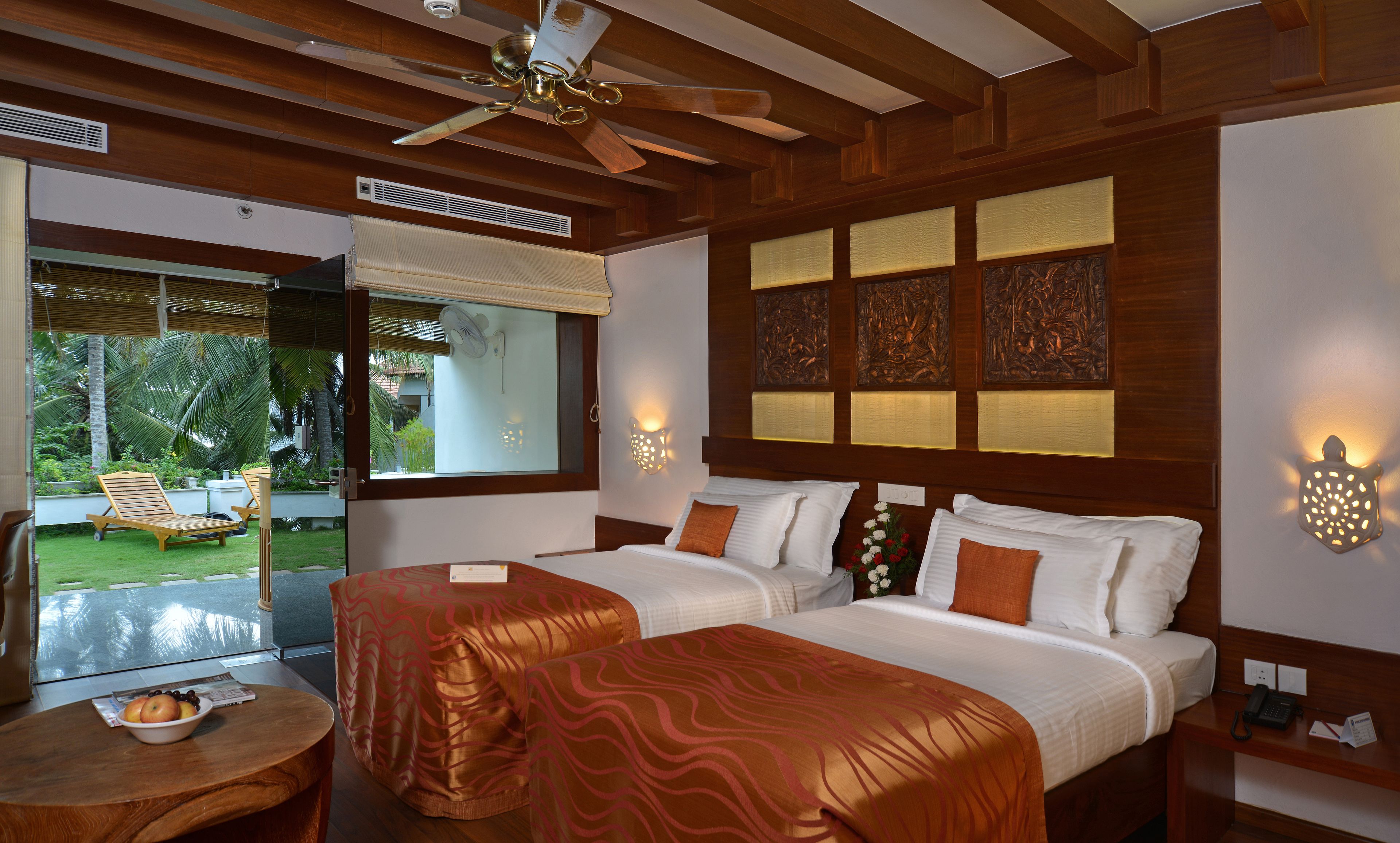 Quarto de hotel com duas camas, um ventilador de teto, uma janela com vista para palmeiras e uma mesa pequena com uma tigela de frutas.
