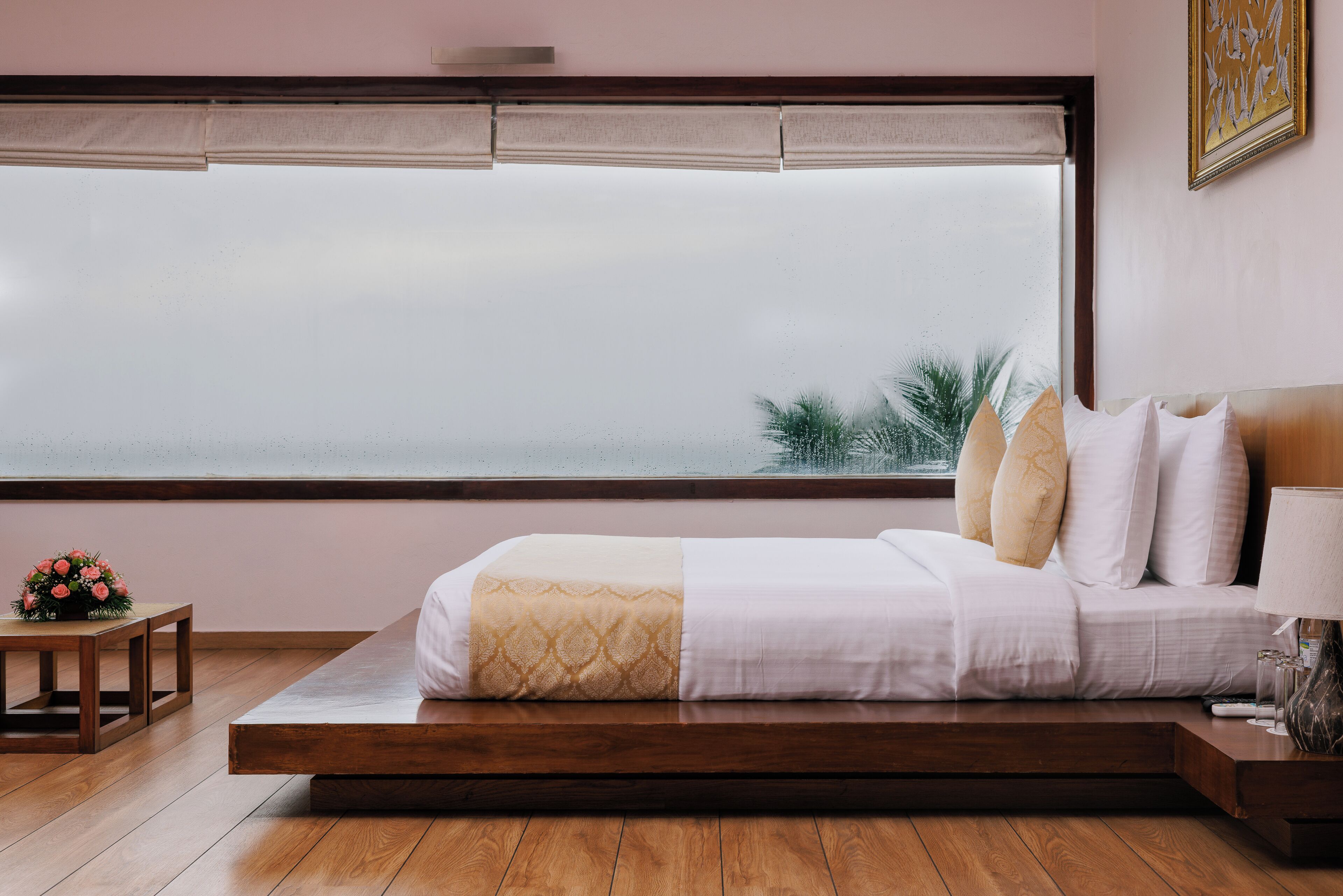 A modern bedroom with a large window, a bed with white and beige bedding, a small wooden table with a flower arrangement, and a nightstand with a lamp.