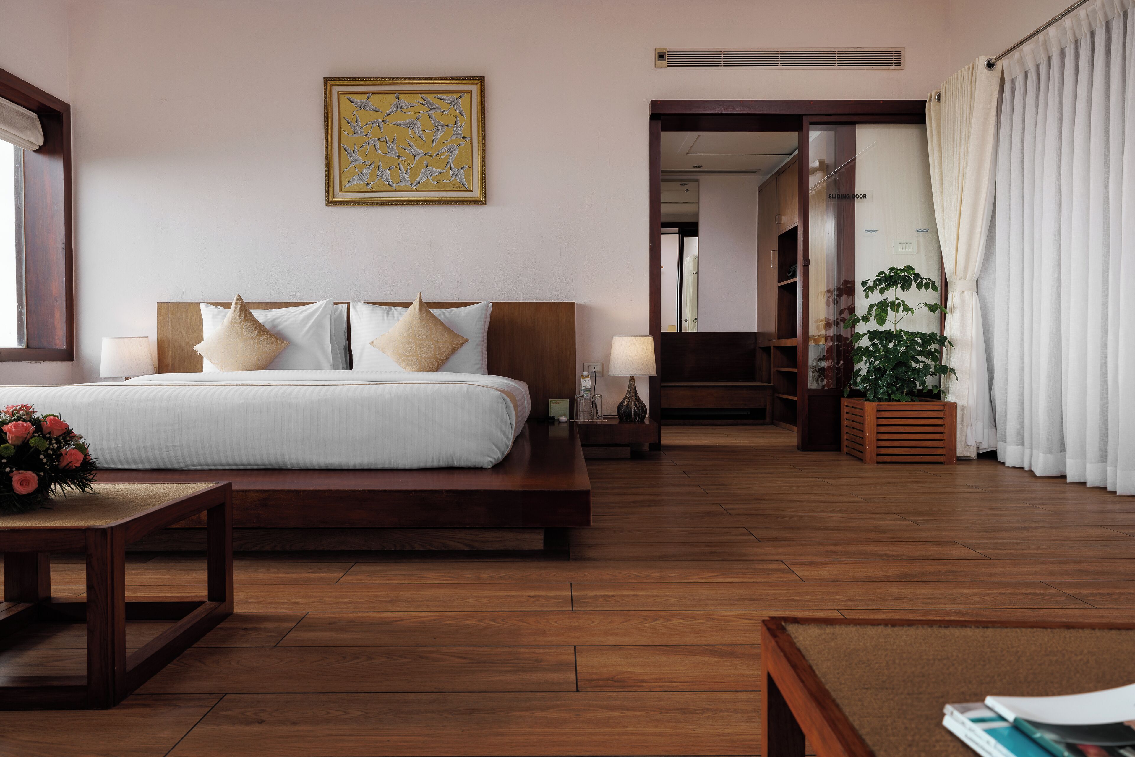 A hotel room with a large bed, bedside tables, a nightstand, a wooden floor, a potted plant, and a framed artwork on the wall.