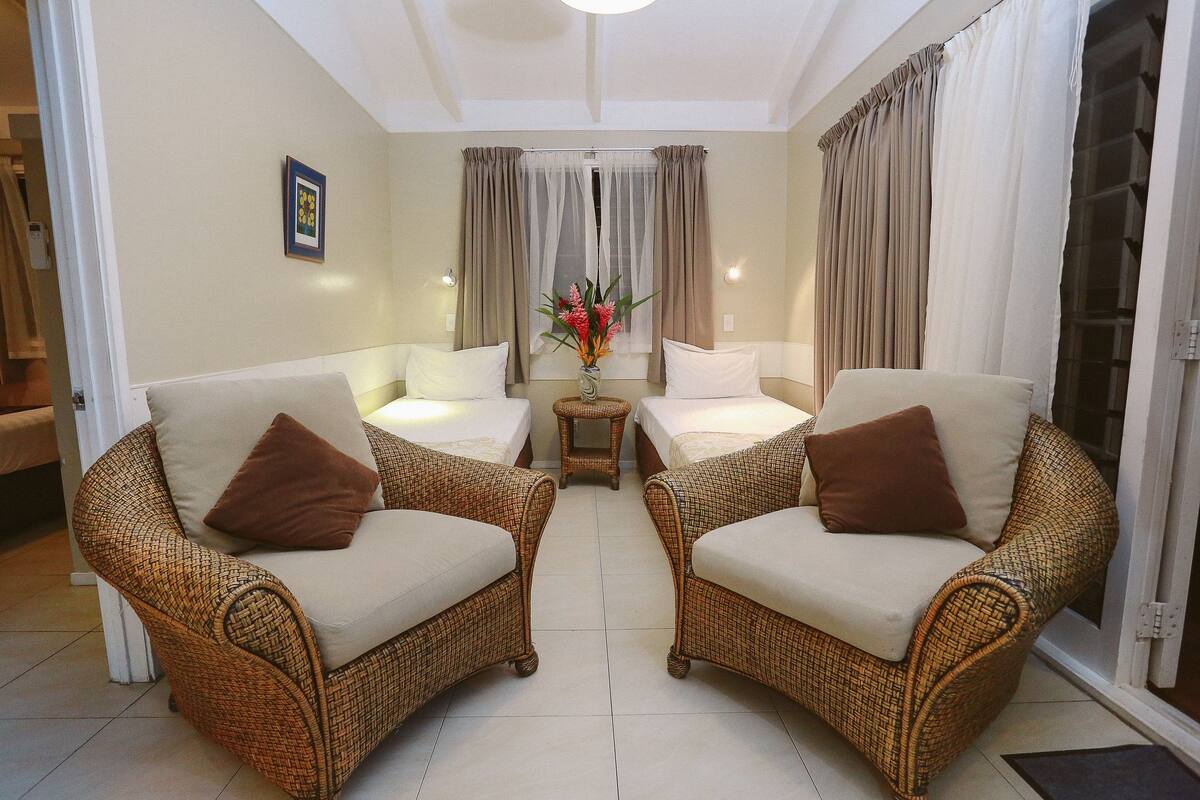 Una habitación con dos sofás de mimbre, una mesita con un jarrón y una cama con cabecero blanco.