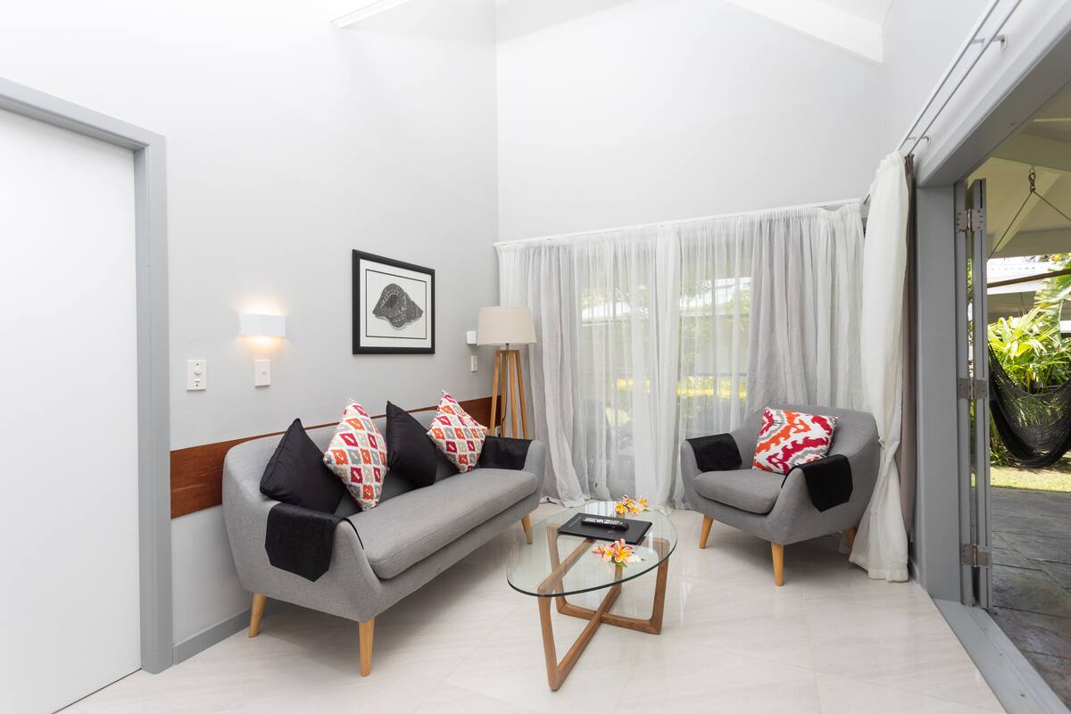 Una sala de estar moderna con un sofá gris, un sillón gris más pequeño, una mesa de centro de vidrio y un ventanal con cortinas traslúcidas.