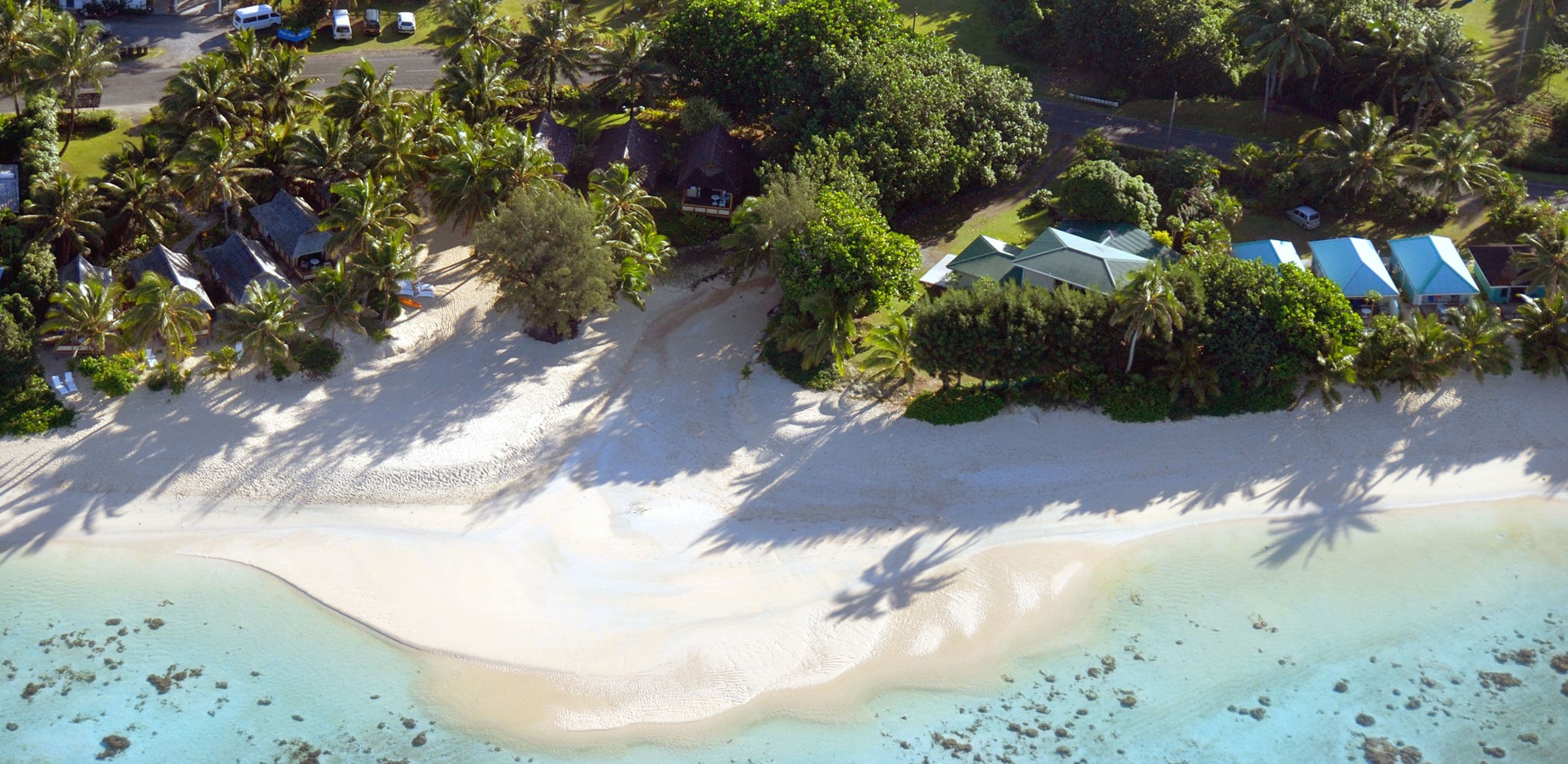 Vista aérea de un complejo turístico en la playa con varias cabañas tipo bungalow, palmeras y una playa de arena.