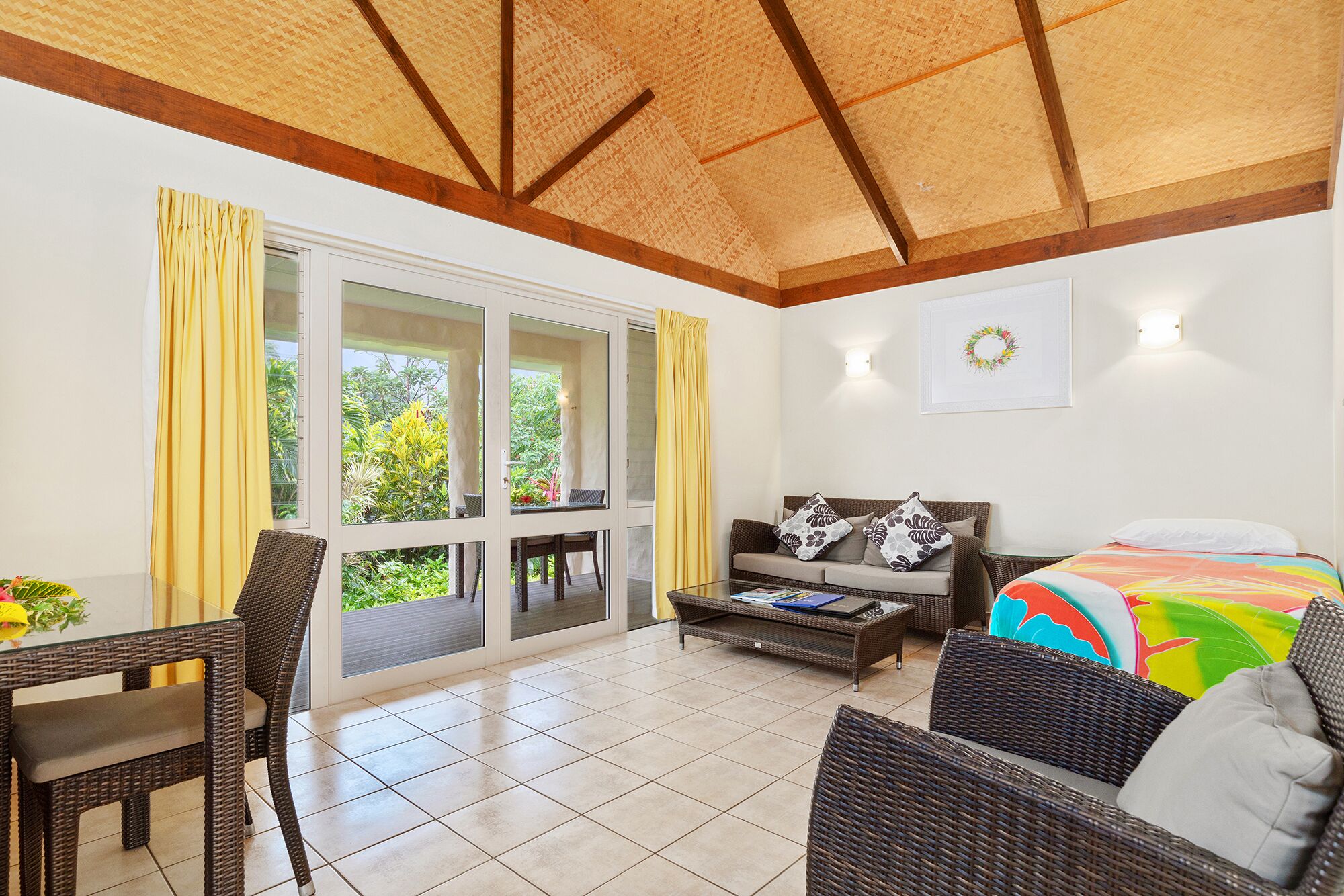 A spacious room with a large sliding glass door leading to a balcony, a bed with a colorful bedspread, wicker furniture, and a wall-mounted light fixture.