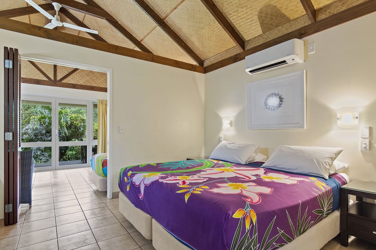Ein Schlafzimmer mit einem großen Bett, einem Deckenventilator, einer Klimaanlage und Blick auf einen Garten durch eine Glastür.