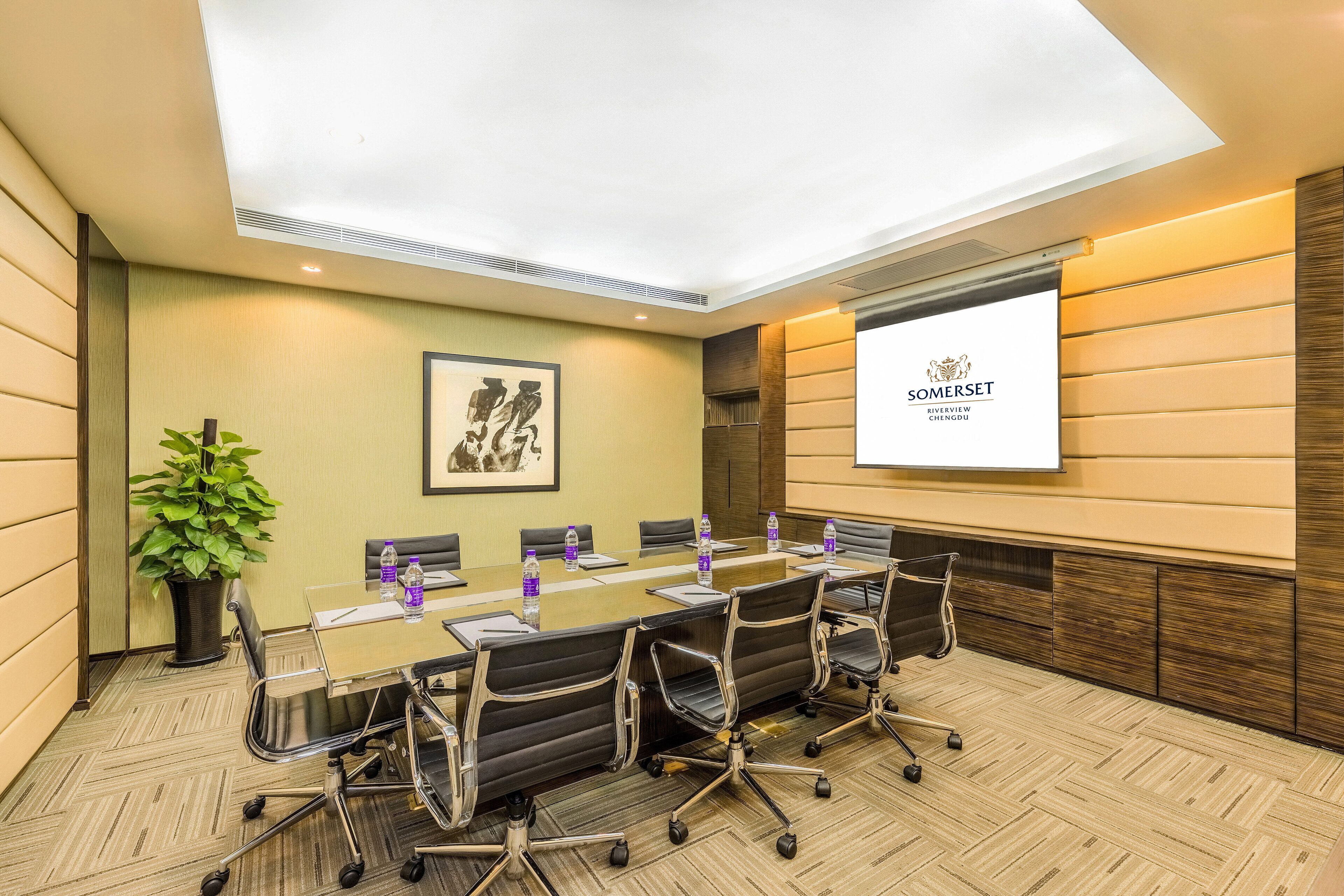 A conference room with a long table, chairs, a projector screen, and a plant.