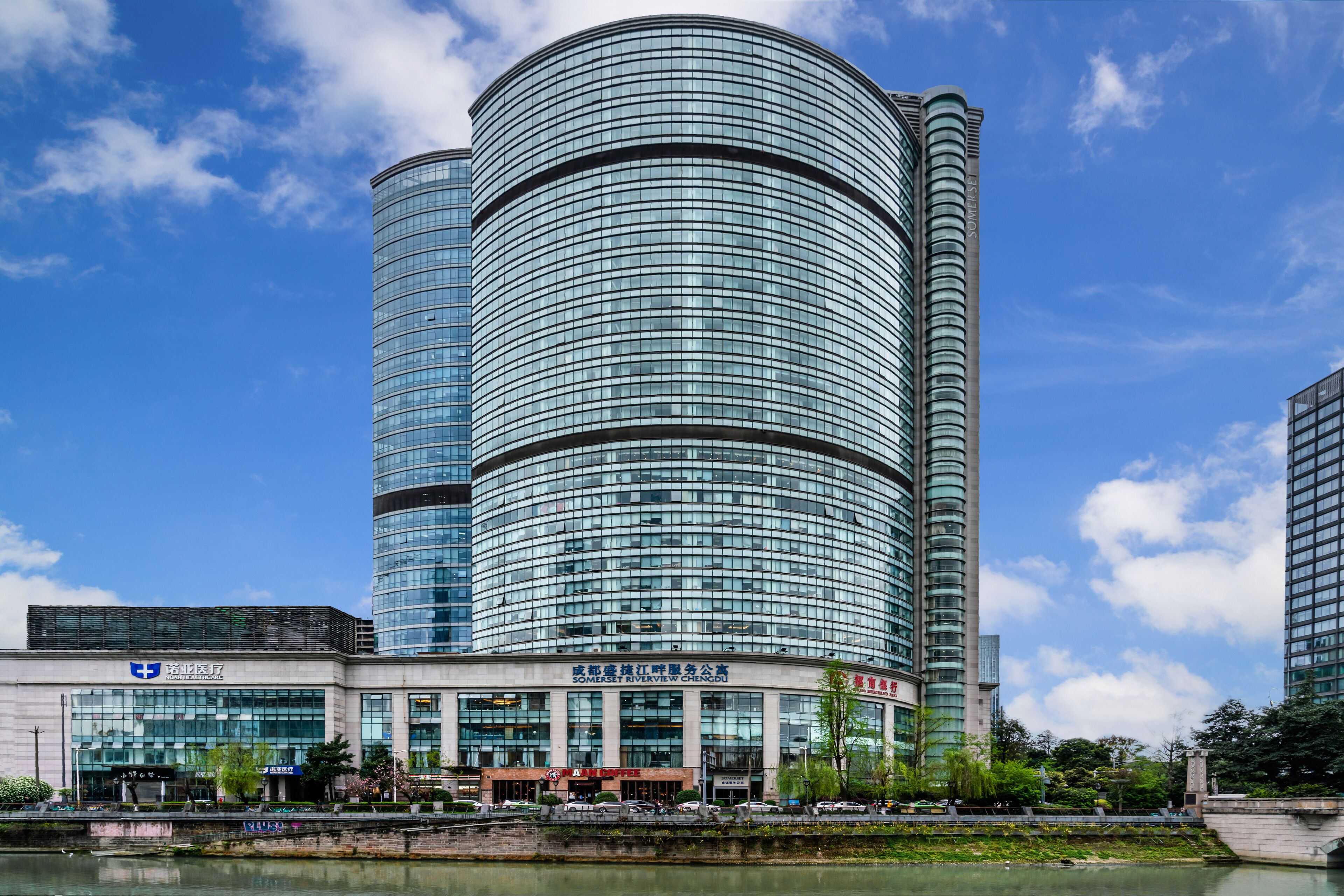En modern byggnad med en böjd glasfasad, en skylt med texten ”Somerset Riverside Chengdu” och en spegelbild i vattnet.