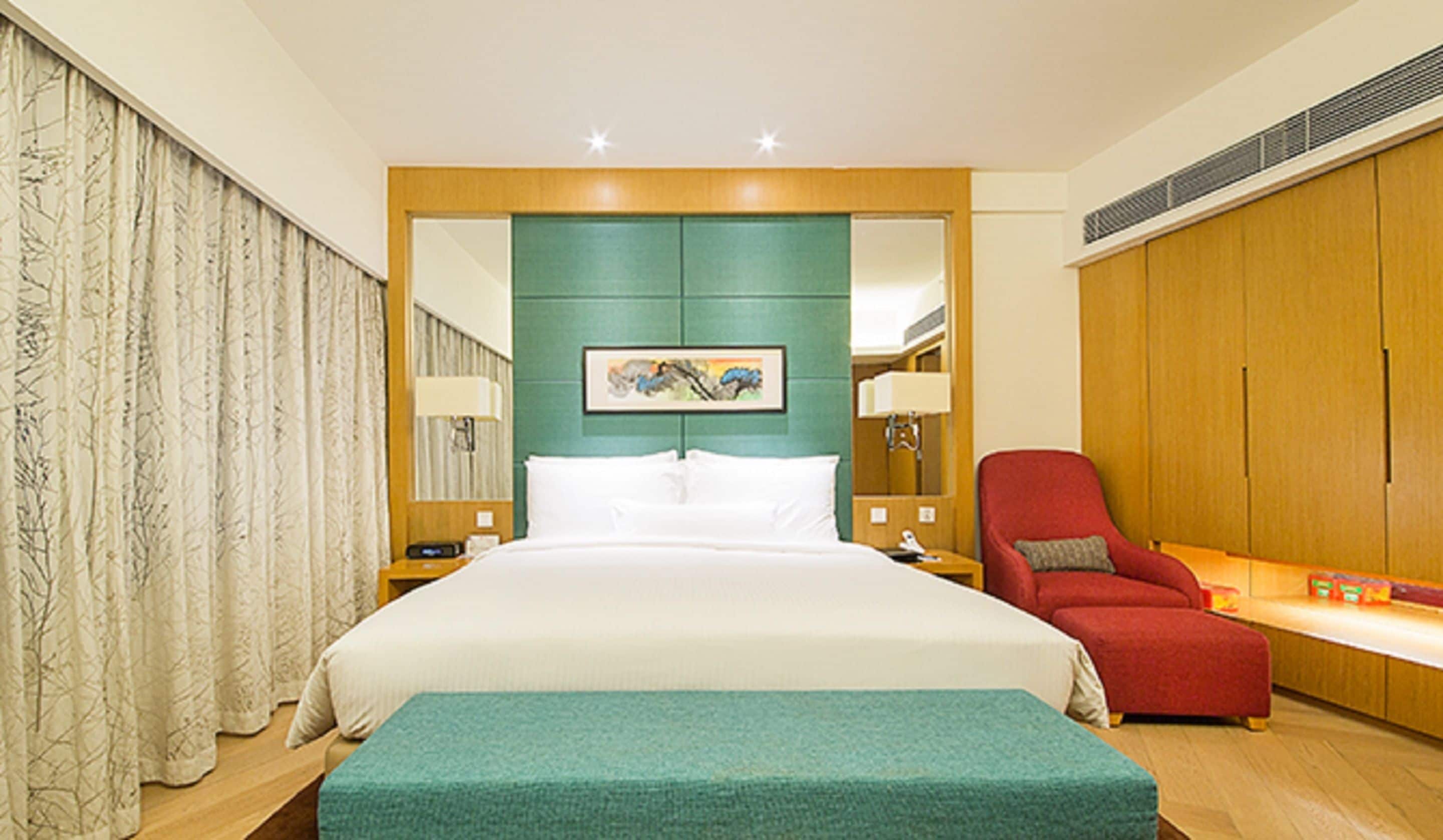 A hotel room with a large bed, a red armchair, a small table with a lamp, and a painting on the wall.