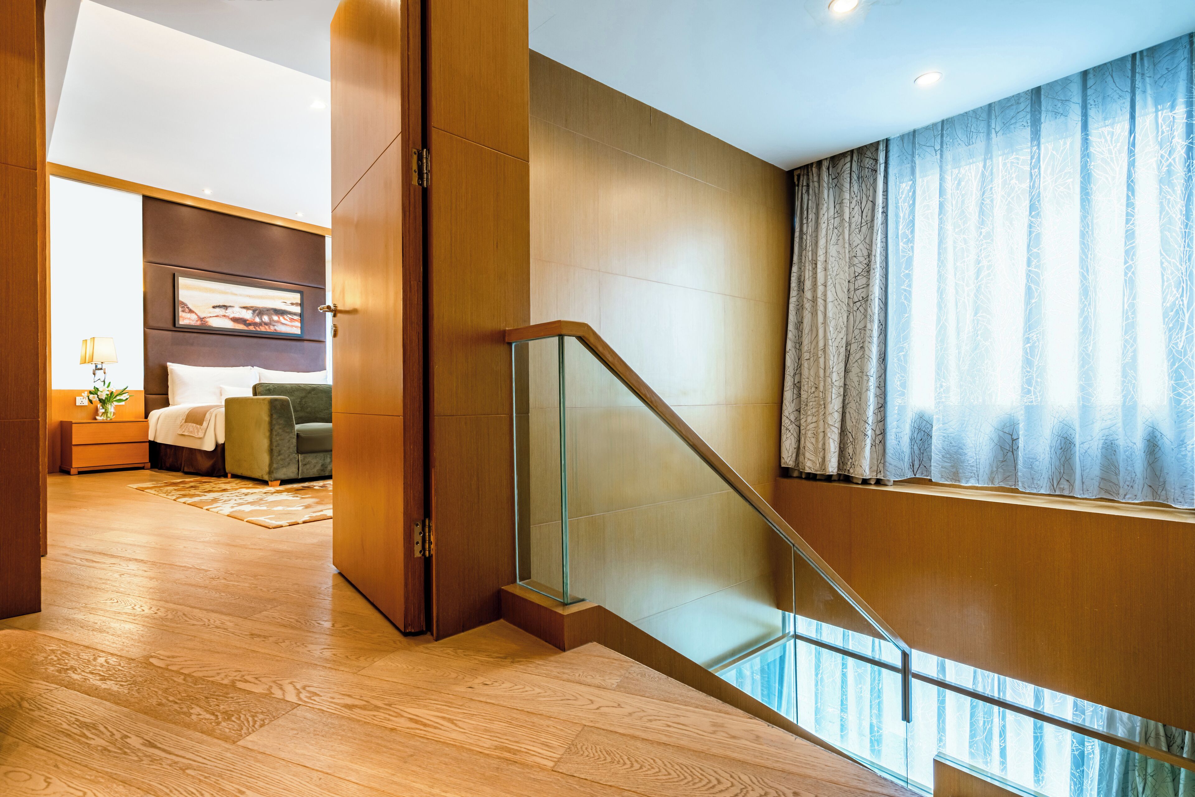 Una habitación de hotel moderna con piso de madera, una escalera de cristal, una cama con una silla verde y un ventanal con cortinas.