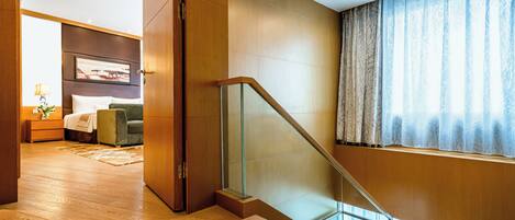 Una habitación de hotel moderna con piso de madera, una escalera de cristal, una cama con una silla verde y un ventanal con cortinas.